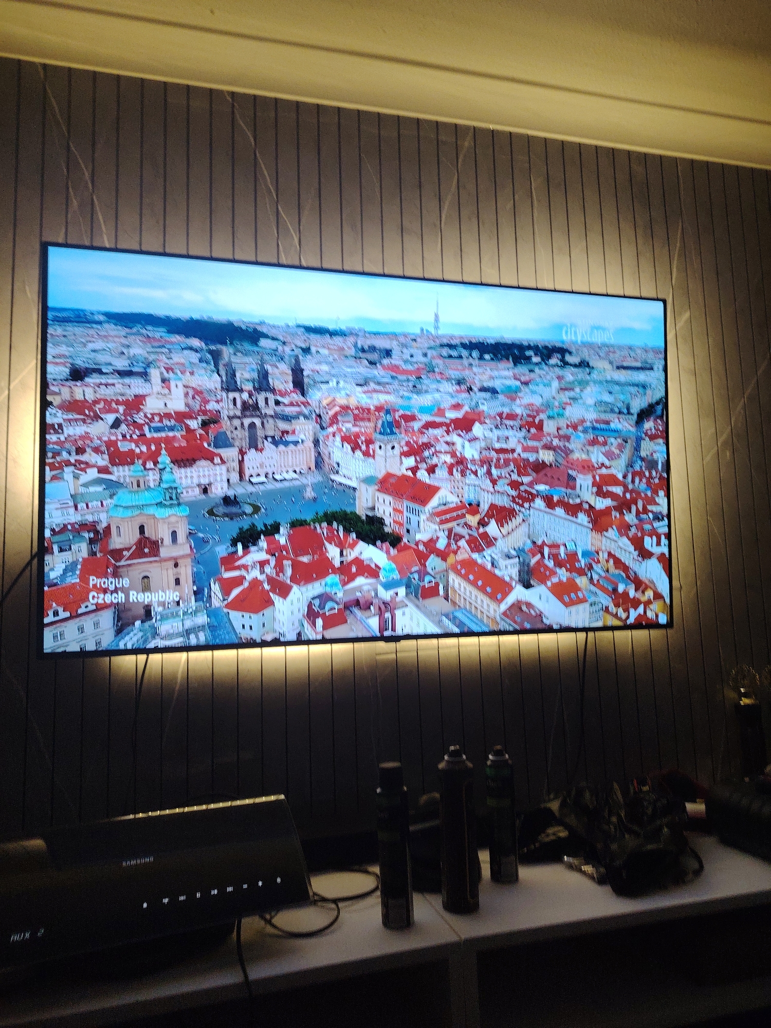 The image displays an indoor scene featuring a large, wall-mounted television screen. The television is displaying an aerial view of a city, clearly identifiable as Prague, Czech Republic, with prominent text overlays "Prague" and "Czech Republic" in the lower-left corner and "cityscapes" in the upper-right corner. The city view predominantly features buildings with numerous red-tiled roofs and several distinct architectural landmarks, including a large square with people and a church with a teal-colored dome.
The television is framed by a vertical panelled wall, dark grey or black in color, with subtle, light-colored, diagonal, thin striations resembling a marble or textured pattern. A strip of warm, yellow backlighting illuminates the wall immediately behind and below the television.
Below the television, on a white surface that appears to be a shelf or media console, sits a black Samsung soundbar. Its front panel features several glowing white indicator lights and "SAMSUNG" branding. Cables are visible near the soundbar. To the right of the soundbar, an assortment of dark objects is present, including several dark cylindrical containers, possibly aerosol cans, and other indistinct dark items like bags or electronics. The overall lighting in the room, apart from the TV screen and its backlighting, is dim. No people are visible in the scene.