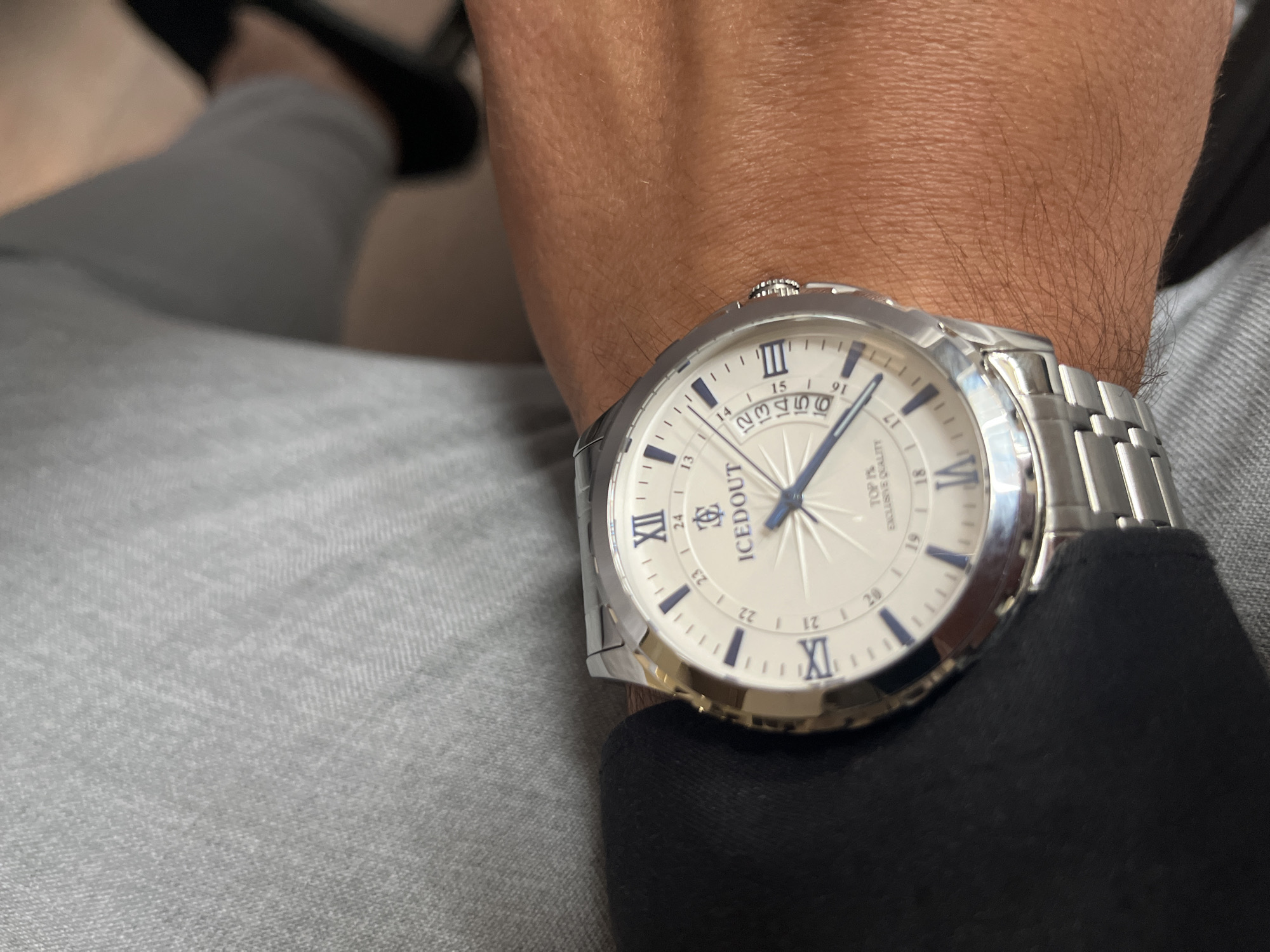 A close-up shot features a man's wrist adorned with a silver "Iced Out" watch. The watch has a white face with blue numerals and hands. The man is wearing a gray pant or jeans, and a black sleeve is visible near the watch.