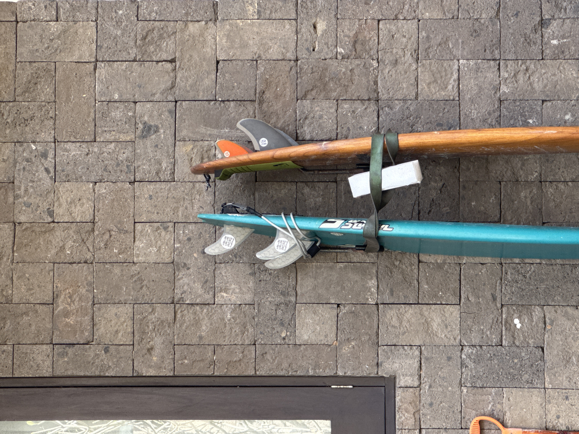 In Kuta Selatan, Indonesia, two surfboards are propped vertically against a textured stone wall. The surfboard on the left is teal and has three gray fins attached. It is secured to the wall by a green strap. The surfboard on the right is made of wood with a green fin attached and an orange fin above it. It is also secured by a green strap. To the left of the surfboards, a dark brown window frame is visible. The stone wall is composed of rectangular blocks with a rough, natural finish. A portion of an orange shovel handle is visible at the bottom left of the frame. The scene appears to be outdoors, possibly in a residential or resort area given the materials and surrounding environment.