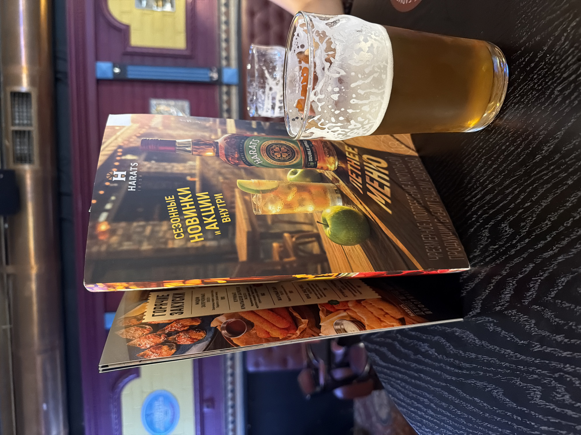 The image shows a close-up of a table in a dimly lit establishment, likely a bar or pub, with two menus and a glass of beer. The menu on the left displays images of fried food, possibly appetizers, with Russian text indicating "Hot Appetizers." The menu on the right features text in Russian such as "Seasonal New Arrivals and Promotions" and "Summer Menu." A bottle of Harat's Irish whiskey is partially visible on this menu, along with a cocktail and an apple. A glass filled with amber-colored beer and a frothy head is prominently placed in the foreground, to the right of the menus. The table surface has a dark, textured wood grain pattern. The background is blurred, but suggests a reddish-brown wall or booth seating and some indistinct architectural details, possibly an air vent. The lighting is warm and atmospheric. There is no specific information in the image to pinpoint the location as Moscow, Russia.