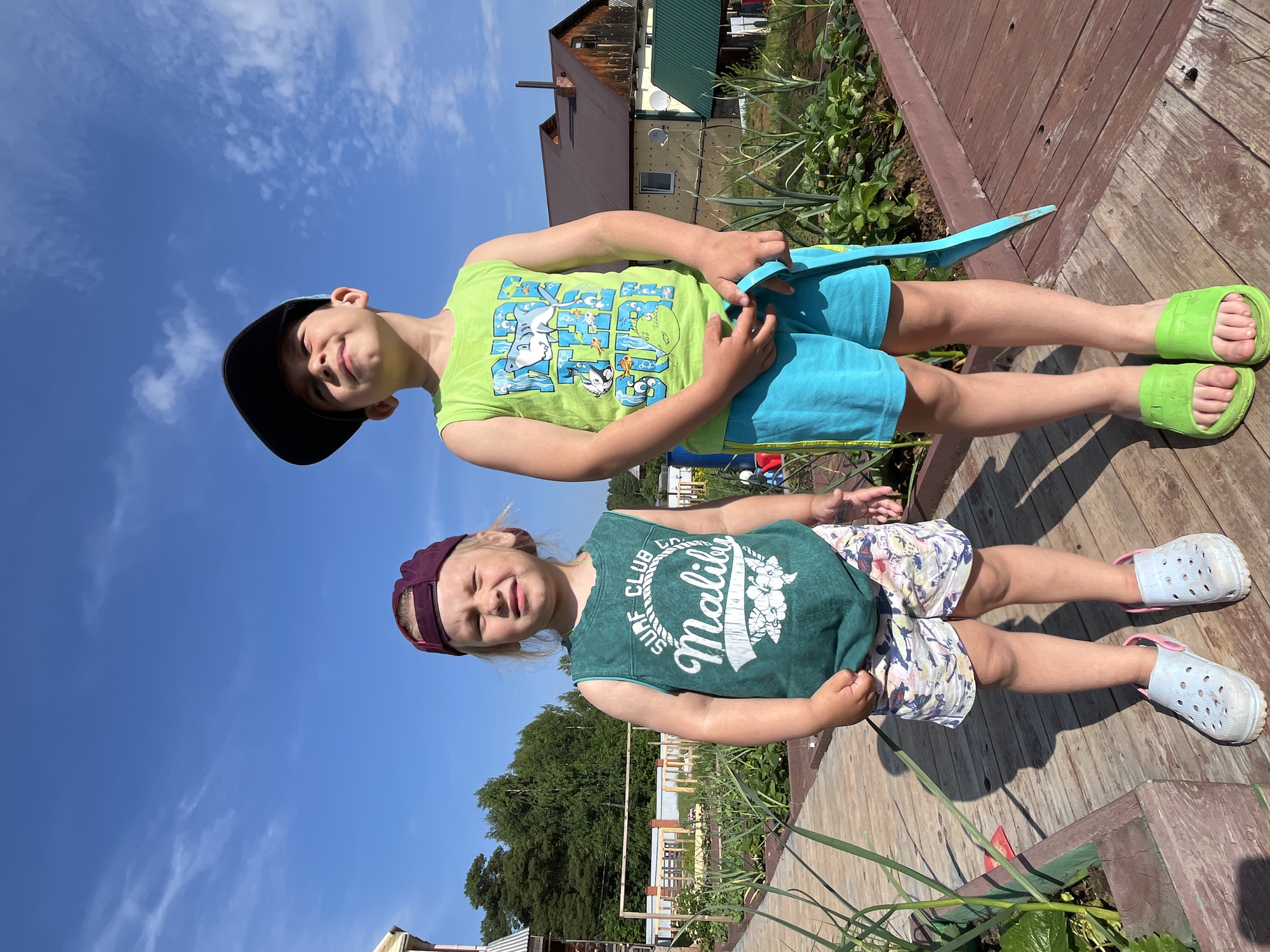 Two children, a young girl and a boy, stand on a wooden pathway. The girl, on the left, wears a dark purple baseball cap, a green tank top with "Malibu" written on it, and patterned shorts. She has light-colored shoes. The boy, on the right, wears a black baseball cap, a green tank top with a shark graphic, and bright blue shorts. He holds a blue object in his right hand and wears green slides. They are in an outdoor setting with a garden to their right, featuring rows of plants. In the background, there are trees and residential buildings. The sky above is blue with scattered clouds. The image appears to have been taken on a sunny day. The location is consistent with a dacha, or country house, in Chaikovsky, Russia.