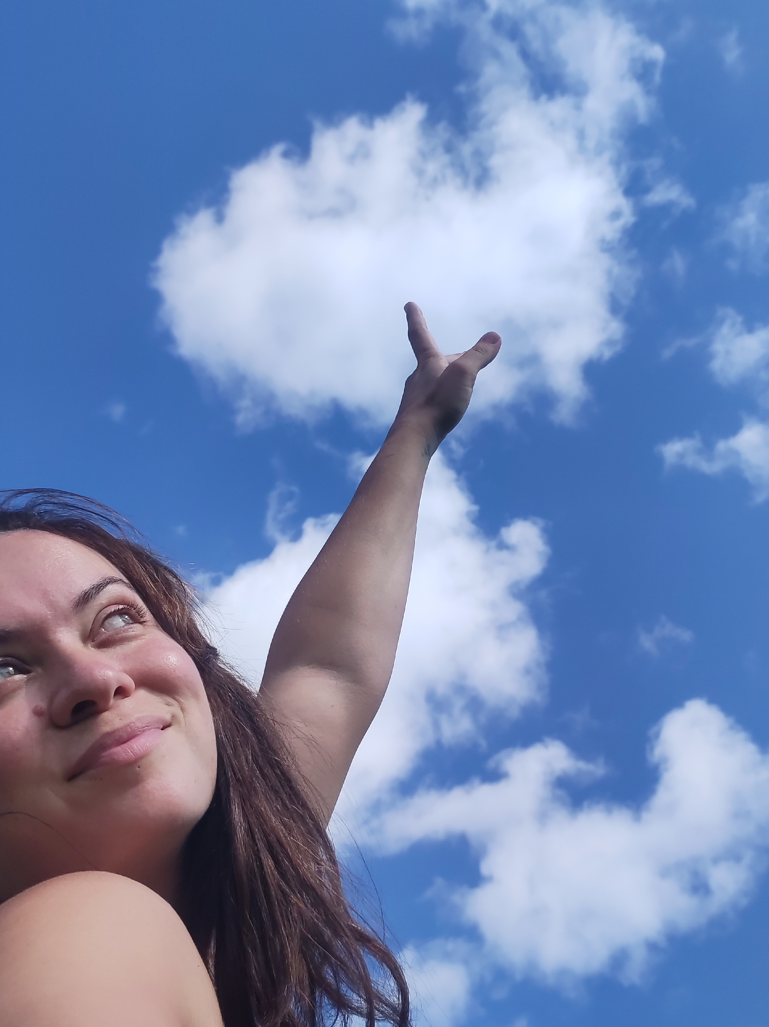 The image shows a young woman looking upwards towards a bright blue sky with fluffy white clouds. Her arm is raised, with her hand outstretched as if reaching for something. She has long, brown hair and a happy expression on her face. The perspective is from a low angle, looking up at the sky and the woman. The image does not provide any information about the location in the city of Фоки, Russia.