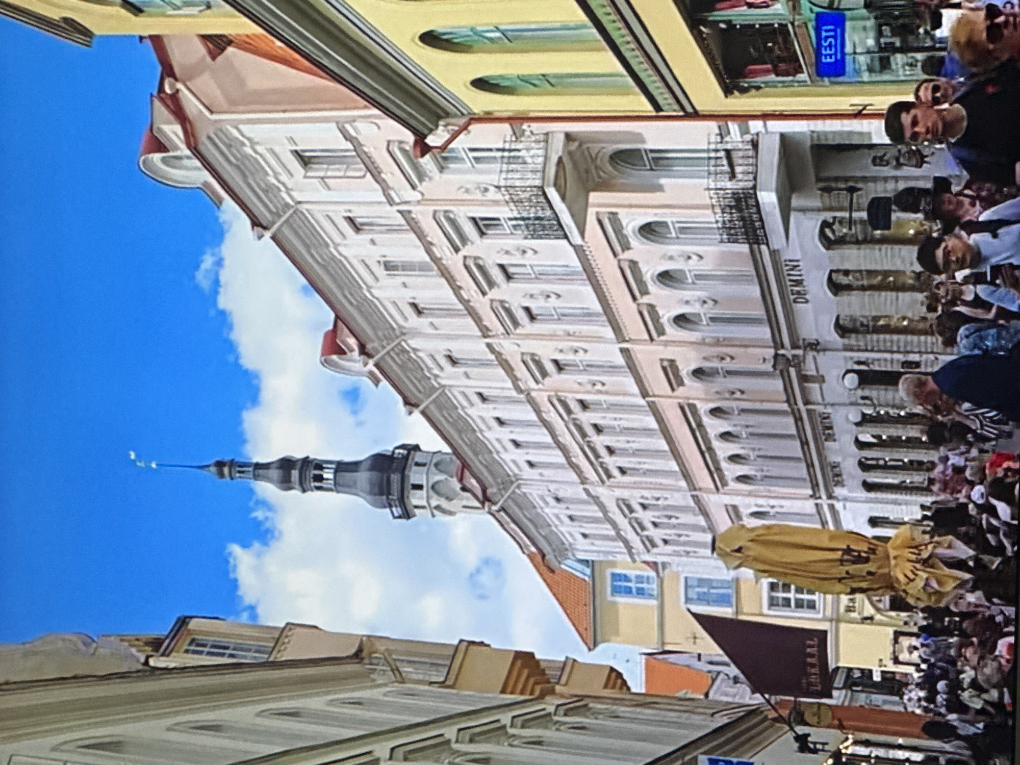 The image depicts a street scene in Tallinn, Estonia, likely in the Old Town due to the historic architecture. Tall, narrow buildings with ornate facades line both sides of the street. A prominent feature is a tall, dark steeple of a church or tower rising into the bright blue sky dotted with white clouds. The buildings are characterized by their pastel colors, white trim, arched windows, and decorative balconies. On the right side of the street, a yellow building with green arched windows is visible. Several people are walking along the street, some are seated at outdoor tables, suggesting a bustling atmosphere. A large, folded yellow umbrella stands prominently in the foreground. The overall impression is that of a charming, historic European city street on a sunny day.