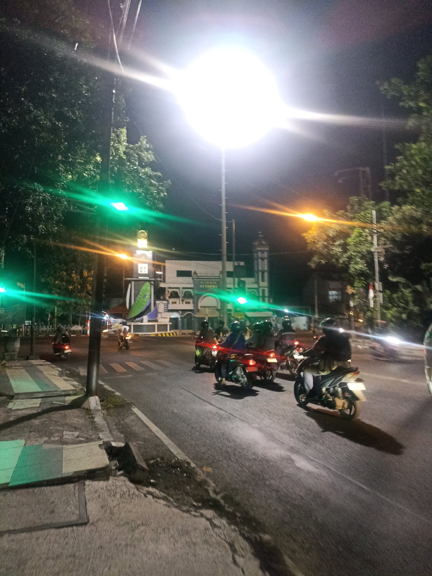 The image depicts a street scene at night in Lamongan, Indonesia, near the Al-Karomah Mosque. A bright street light dominates the upper portion of the frame, casting a harsh glare and lens flare across the sky. In the foreground, a road is visible with several motorcycles and their riders. Some motorcycles are in motion, while others appear to be stopped at an intersection, indicated by a pedestrian crossing. To the left of the road, a sidewalk is present with green and gray colored paving stones. A traffic light displays a green signal. In the background, the Al-Karomah Mosque, a white building with a tall minaret, is visible. Trees line the sides of the road, illuminated by the artificial lights. The overall atmosphere is that of a busy urban street at night.