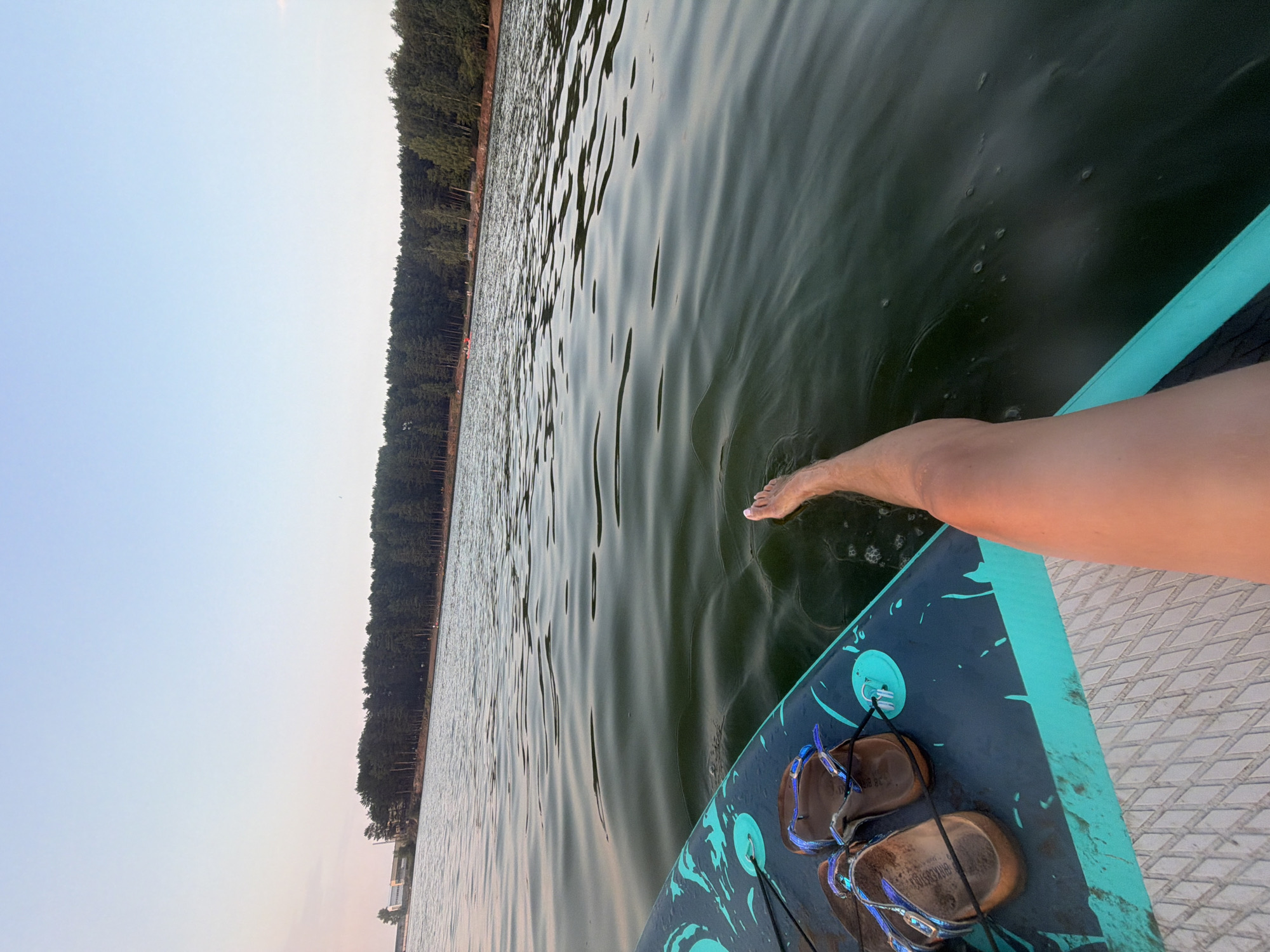 The image shows a person's legs and feet on a paddleboard in a body of water. The paddleboard has a turquoise and black design, and a pair of brown sandals are placed on the front of it. The water is calm with gentle ripples. In the background, a dense forest lines the shore, and a building complex can be seen to the left. The sky is a pale blue, suggesting it is either early morning or late evening. This scene is consistent with the location at the Kama River reservoir in Chaikovsky, Russia, known for its recreational activities.