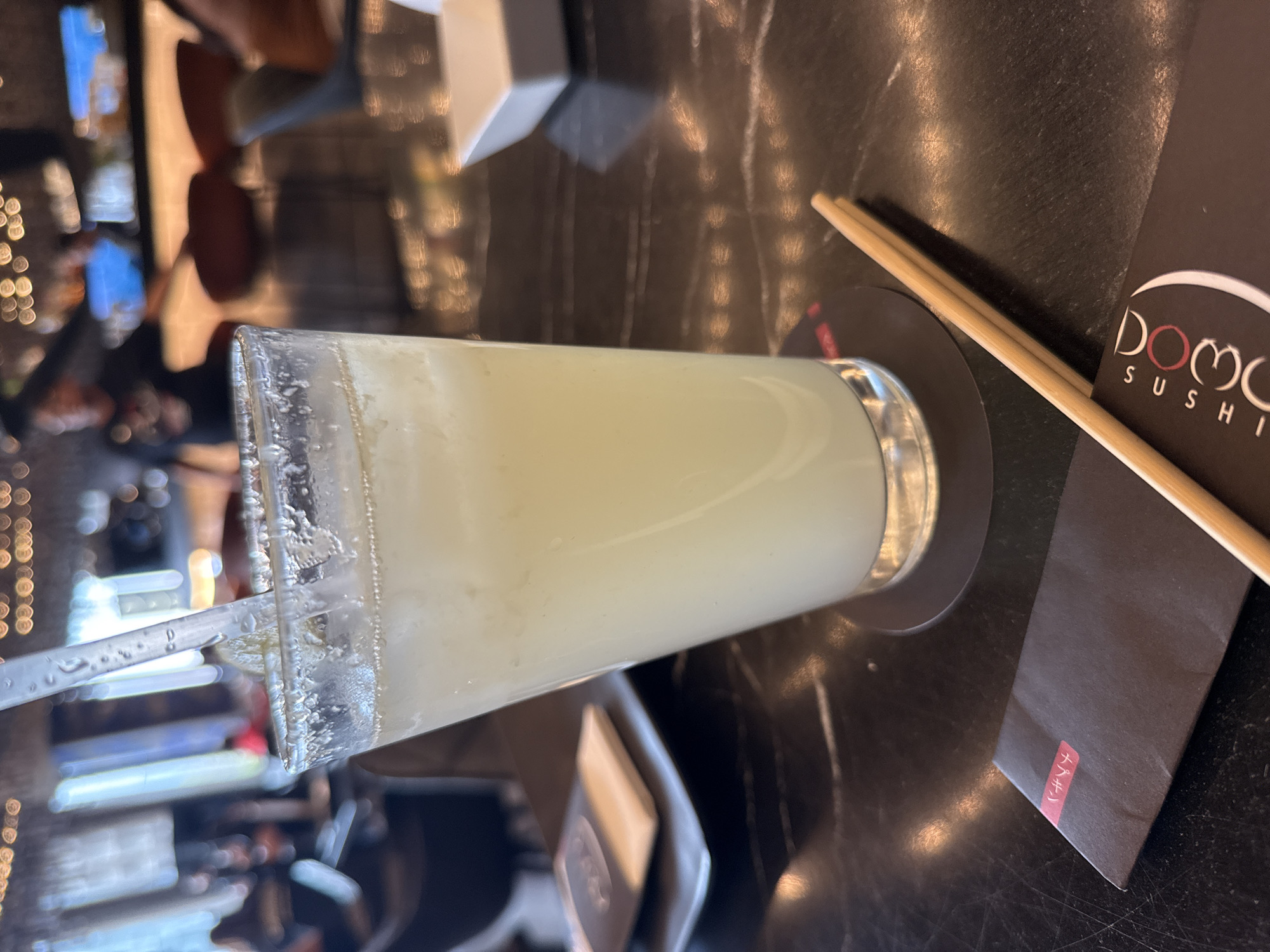 A tilted, eye-level indoor shot shows a beverage glass, likely a margarita, on a black marble-like surface with a Domo Sushi restaurant chopstick holder and chopsticks. The glass has a sugared rim and a plastic straw inside the pale-colored drink. A napkin sits partially under the glass, and a menu with the "Domo" sushi logo is to the right of the drink, partly obscuring the edge of a dark coaster. The background displays blurred lights and suggests the inside of the restaurant, presumably Domo Sushi in Puebla, Mexico. The scene is focused on the drink and sushi-related items, implying a dining setting.