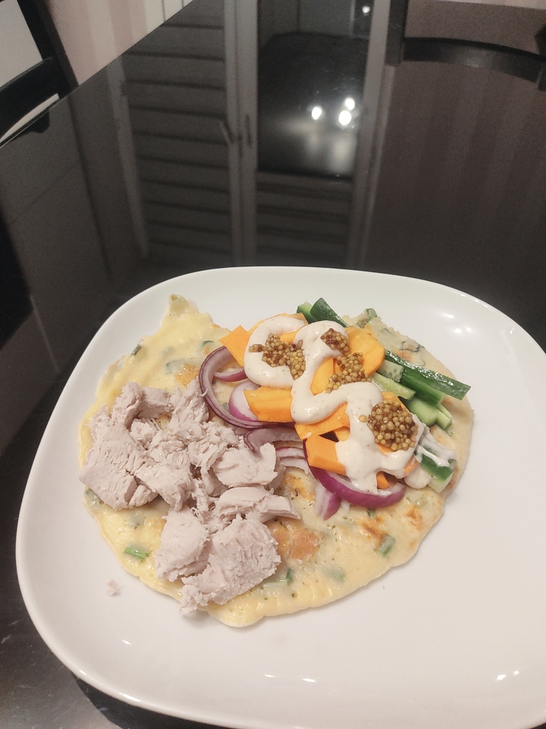 A close-up shot presents a white square plate, upon which rests a culinary creation. The meal consists of a pale yellow base, possibly an omelet or flatbread, topped with assorted ingredients. There is a portion of shredded poultry, likely chicken or turkey, positioned on the left side of the dish. Sliced red onion rings, chunks of orange carrots, and slices of green cucumber are also present. A light-colored sauce, garnished with what appears to be whole grain mustard seeds, is drizzled over the vegetables. The plate is situated on a dark, reflective surface, possibly a dining table. The background shows a blurred view of what seems to be a white closet door. The location is unknown.