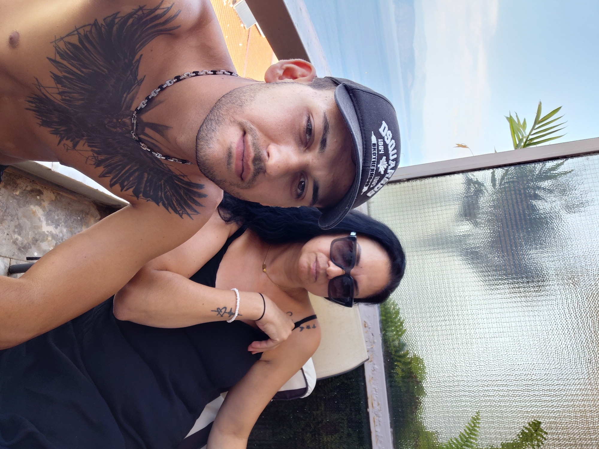 Two individuals are posing for a selfie outdoors, with a body of water and distant hills visible in the background. The man in the foreground, wearing a black baseball cap with a white logo and a silver chain necklace, is shirtless and displays a large black bird tattoo on his chest. To his right, a woman with long dark hair, wearing sunglasses, a black tank top, and bracelets, is seated. The scene appears to be a balcony or terrace overlooking a coastal landscape, likely in the Municipal Unit of Aegio, Greece, suggested by the distinctive scenery. The overall atmosphere is casual, possibly during a vacation or leisure outing.