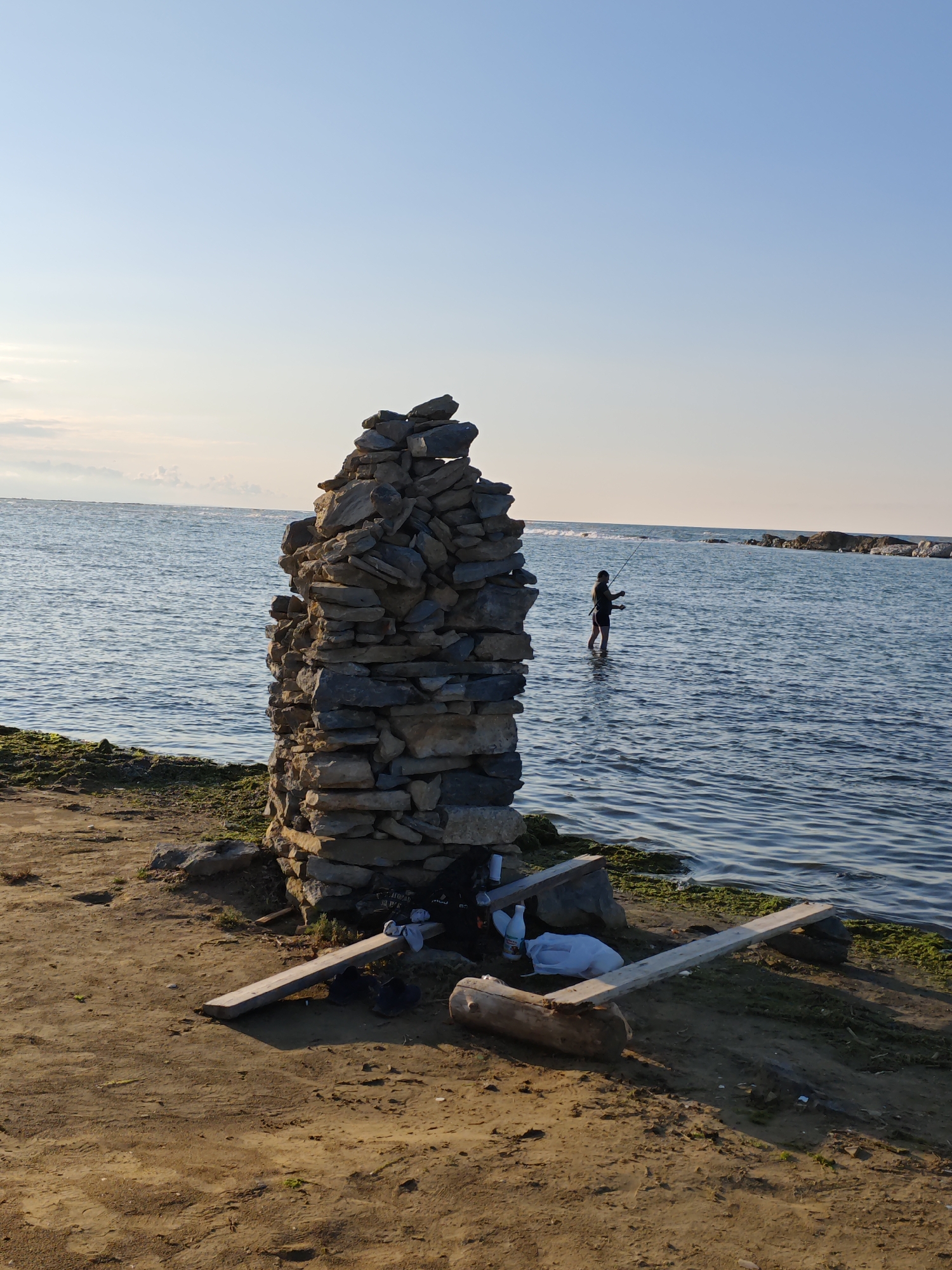 A tall, narrow cairn constructed from stacked flat stones stands on a sandy, rocky shore. The cairn is located on the left side of the image, adjacent to the water. To the right of the cairn, a person is wading in the calm blue water, holding a fishing rod. The water extends to the horizon, with a clear sky above. In the foreground, to the right of the cairn, are two wooden planks, a log, and some plastic bags. The scene suggests a peaceful outdoor setting, likely near a body of water. The location is identified as Izberbash, Russia.