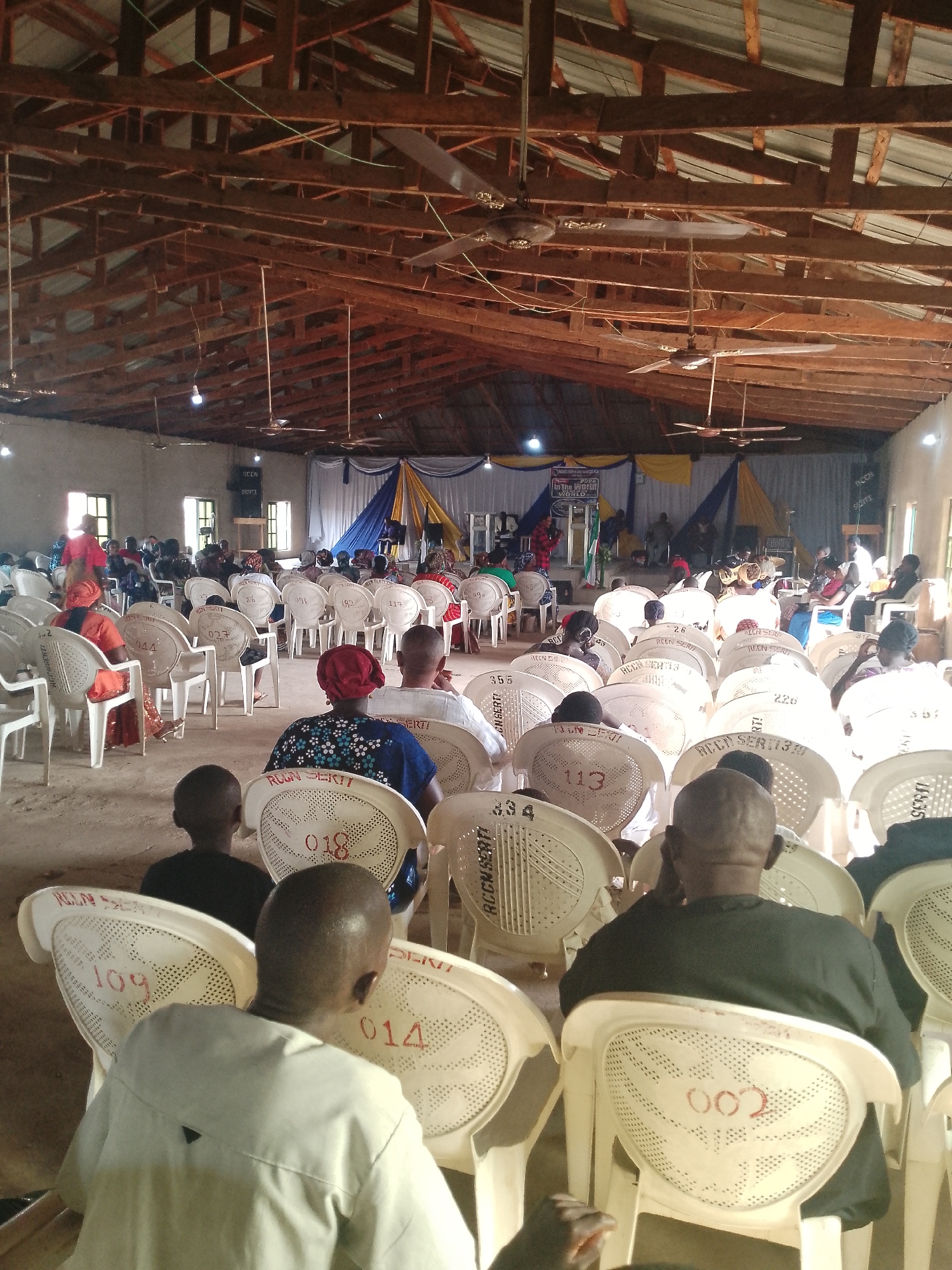 The image depicts an indoor gathering, likely a church service, with a congregation seated in rows of white plastic chairs. The room has a high, wooden-beamed ceiling with exposed rafters and multiple ceiling fans. The walls are light-colored, and there are windows on the left side of the image. The front of the room features a stage area with draped blue and yellow fabric, a podium, and sound equipment. Several people are seated, facing the stage. Some chairs have numbers written on them in red. The overall atmosphere appears to be calm and attentive. The exact location is unknown.