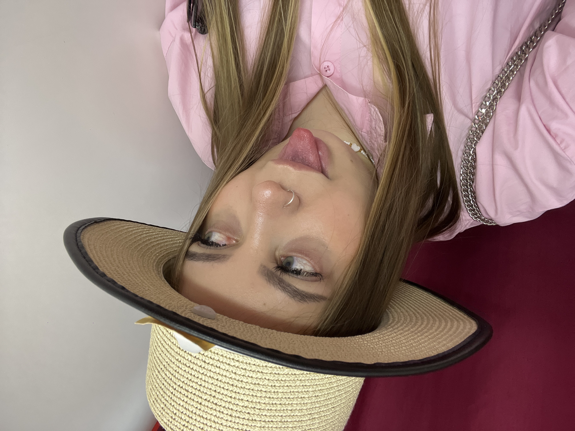 A woman wearing a straw hat and a pink shirt is sticking her tongue out. She has a nose ring and is wearing a necklace. She has long blonde hair. The background is a white wall and a red surface. The image is rotated. There is no information indicating that this image was taken in Novopokrovskaya, Russia.