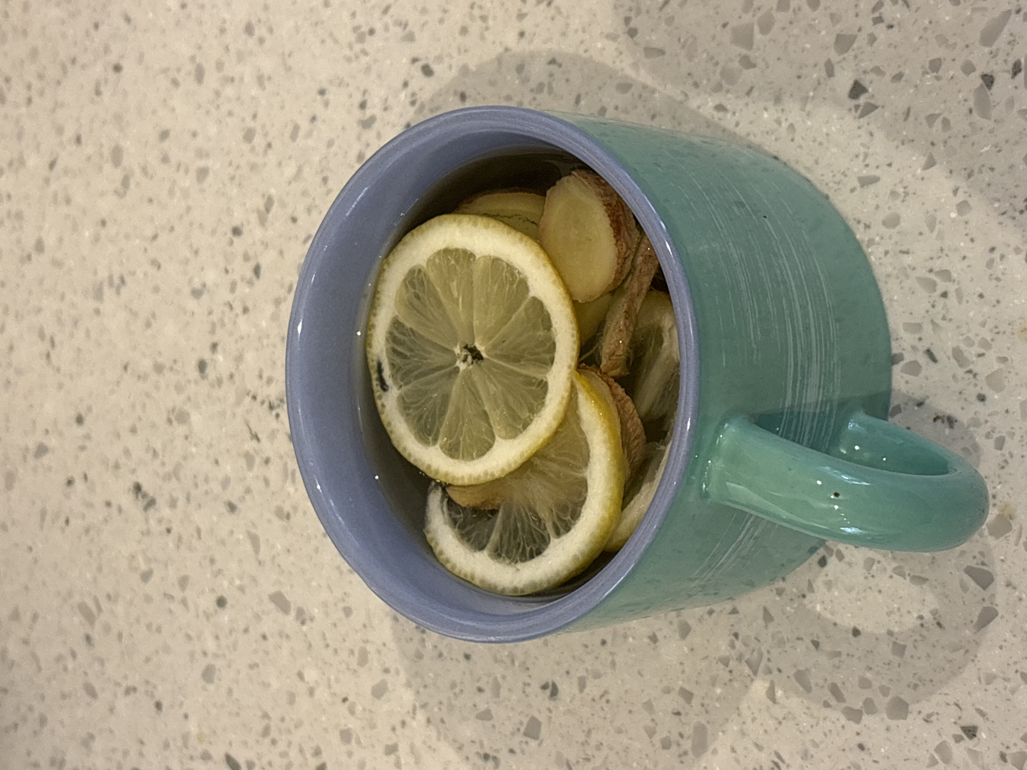 An overhead, slightly angled shot captures a light blue-teal ceramic mug resting on a speckled countertop in a domestic setting in London, United Kingdom. The mug's interior rim is a lighter, possibly lavender-blue. It is filled with a clear liquid, prominently featuring several thin, circular slices of fresh lemon and irregularly shaped pieces of sliced ginger. The bright yellow lemon slices, with their white pith and translucent pulp, float on the surface, while the pale ginger pieces are visible among them, some partially submerged. The contents suggest a hot beverage like lemon-ginger tea or an infusion.
The countertop beneath the mug is a light, possibly off-white or cream color, patterned with numerous small, irregular grey and dark specks, consistent with a quartz or granite surface. The lighting appears to come from the upper left, casting a soft shadow of the mug and its handle onto the countertop, visible to the right and below the mug. No people are visible, and there is no discernible text on any object. The image focuses entirely on the mug and its contents, conveying a moment of preparing or enjoying a warm, healthful drink.