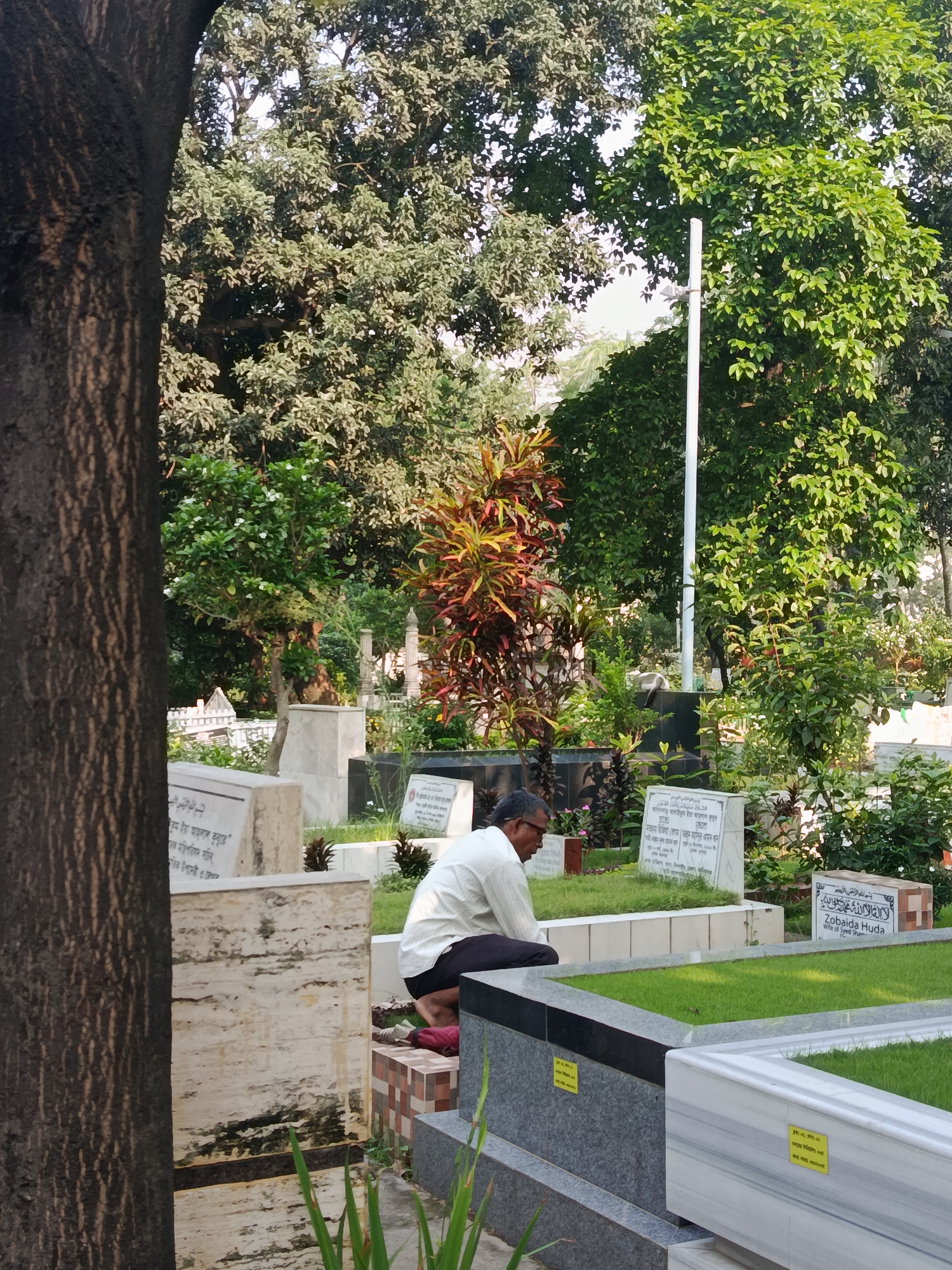 A man wearing a light-colored shirt and dark pants is kneeling in a graveyard. The gravestones are made of marble and granite, some inscribed in Arabic. In the background, there are trees and plants. The location is presumed to be in Dhaka, Bangladesh, based on the OCR text on the gravestones.
