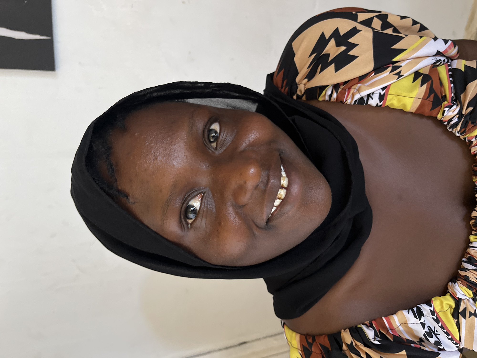 A woman smiles at the camera. She is wearing a black head covering and a colorful patterned top. The location appears to be indoors, with a white wall visible in the background. A dark object is partially visible on the wall. The city and specific location are unknown.