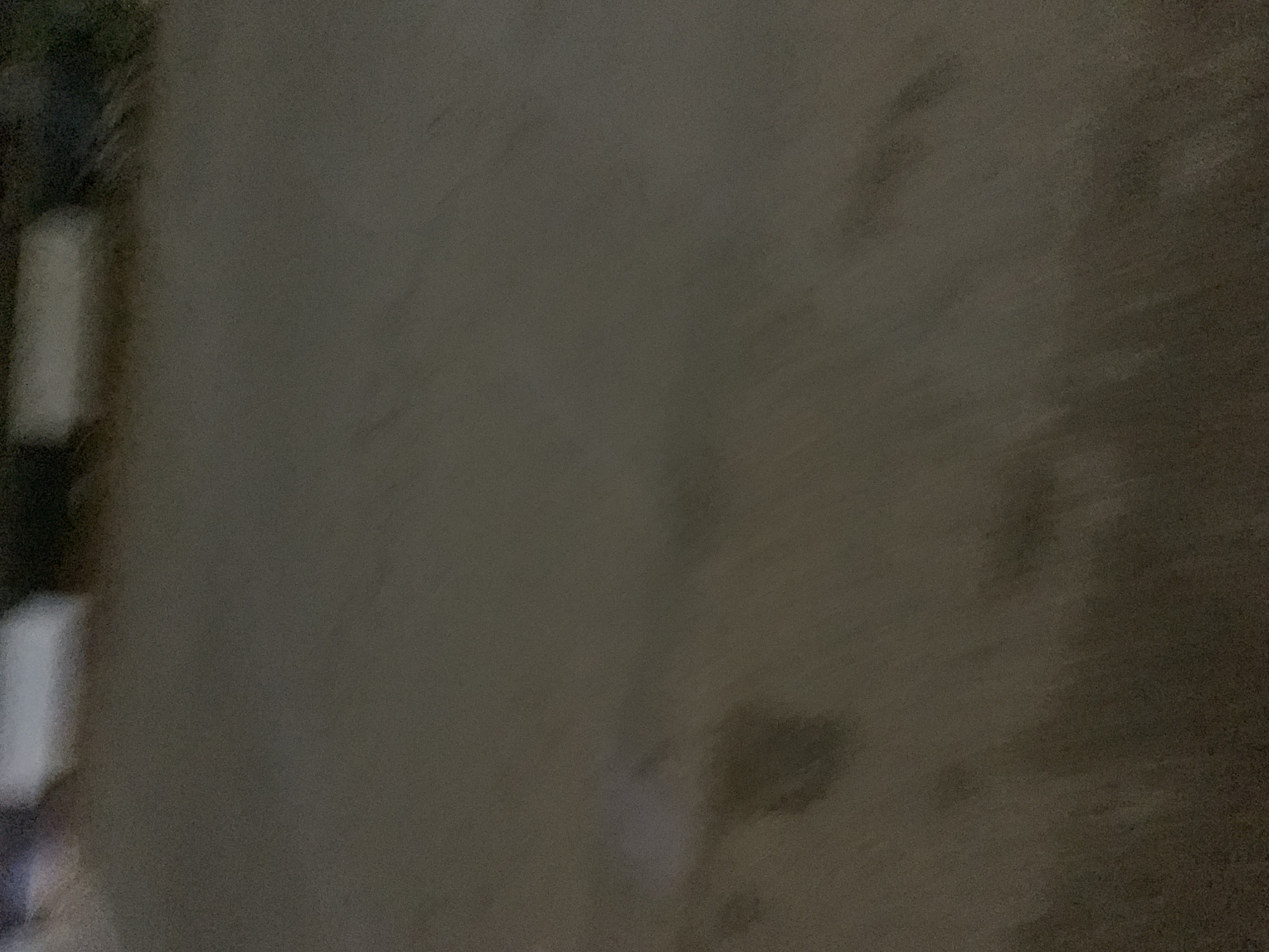 The image is an out-of-focus, low-light shot of a dark, possibly wet, surface. There are indistinct shapes and patches of lighter and darker tones, suggesting variations in texture or the presence of objects. The upper portion of the image shows blurred horizontal bands of lighter colors against a darker background, which could be structures or lights. The overall impression is of an outdoor environment at night or in dim conditions, with no clear subjects or actions discernible.