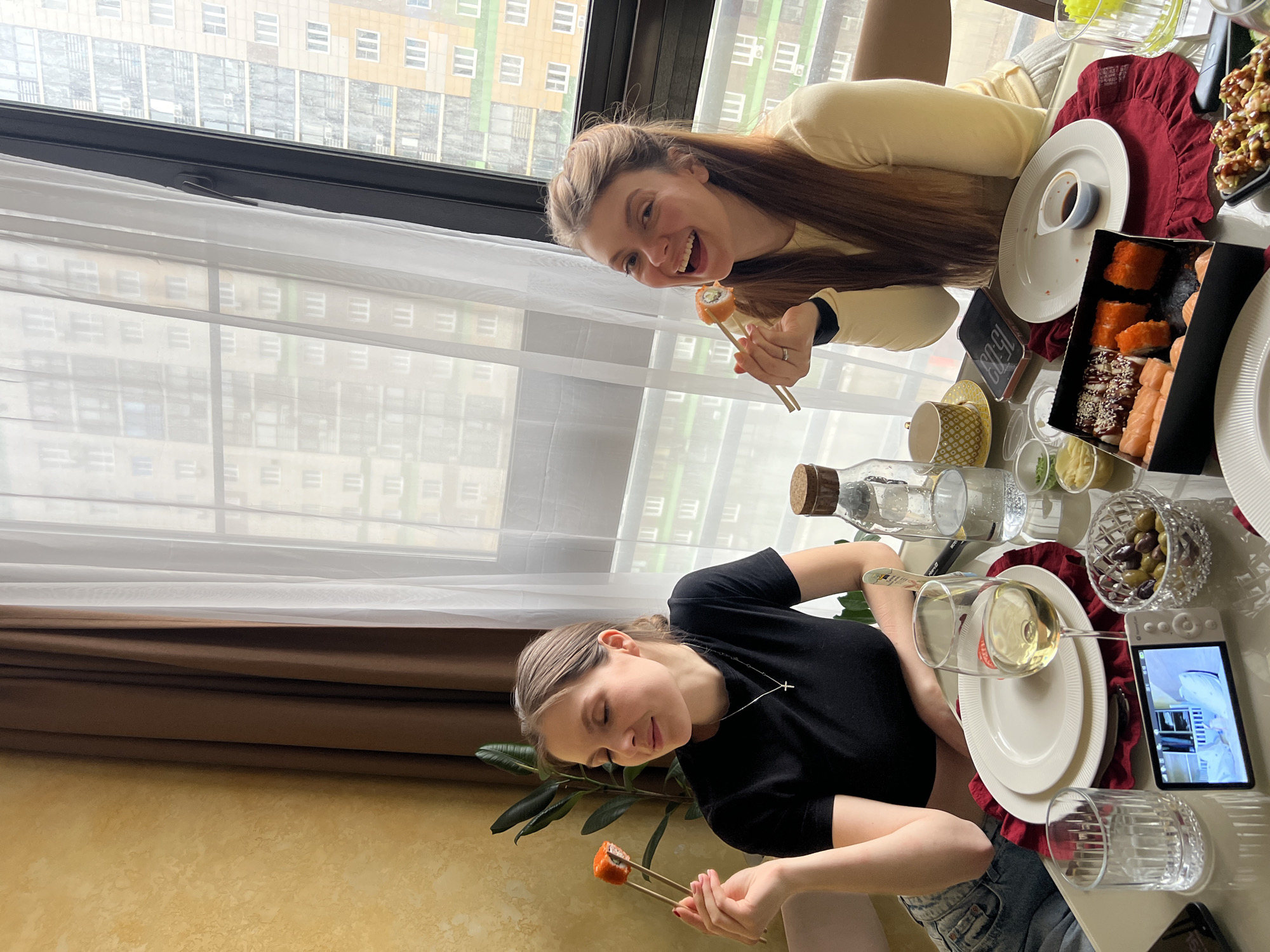Two young women are dining together in an apartment, likely in Khimki, Russia. The location is suggested by the building visible through the window. One woman, wearing a yellow shirt and with long brown hair, is holding a piece of sushi with chopsticks, smiling towards the camera. The second woman, wearing a black shirt, is also holding sushi with chopsticks. They are seated at a table covered with various dishes, including sushi, olives, sauces, and drinks. A tablet displaying a video is also on the table. The background shows a high-rise building with multiple windows and a light yellow and green exterior. A window with white curtains and brown drapes is visible behind the table.
