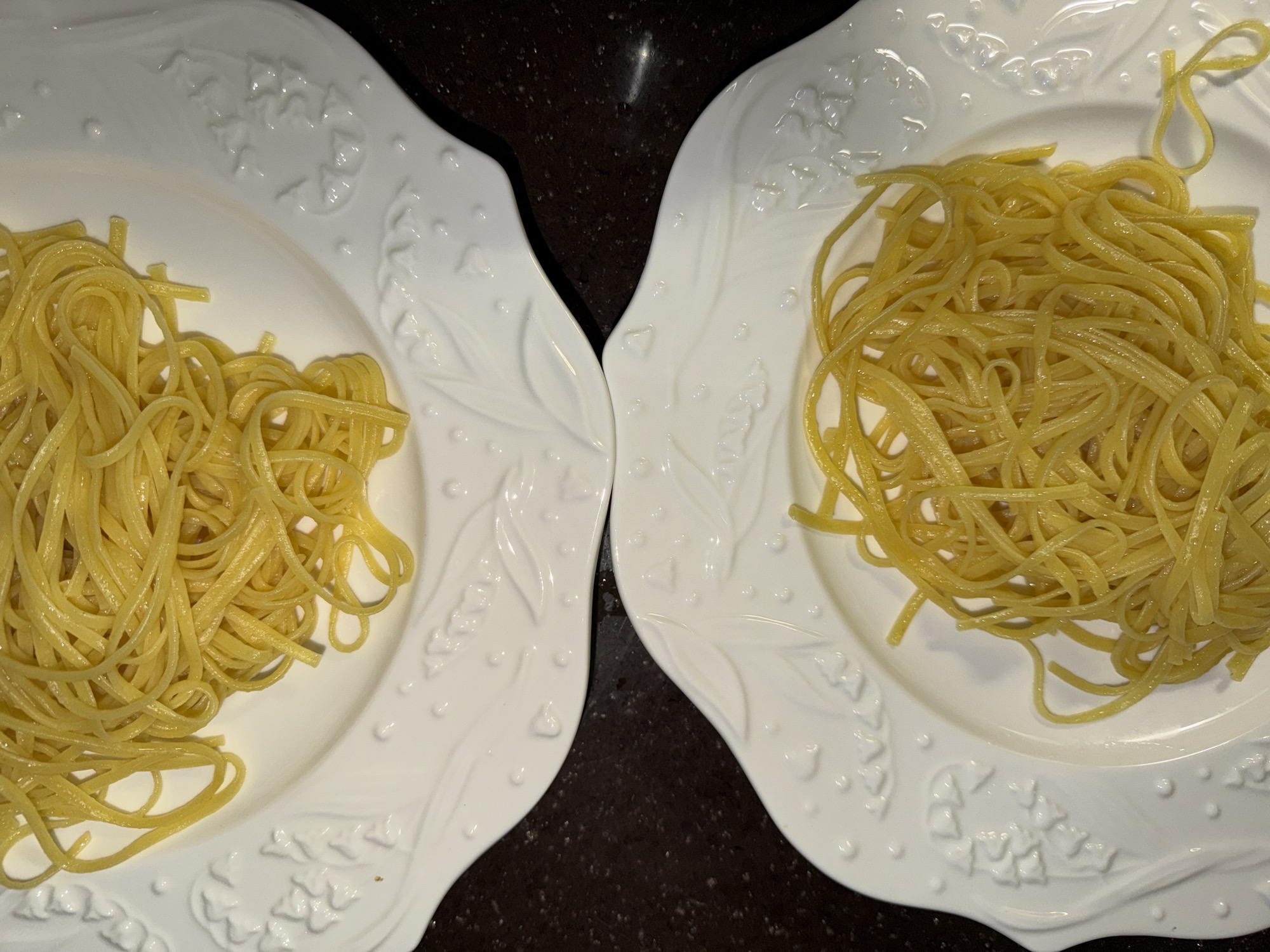 Two white, intricately designed plates, each holding a serving of plain, cooked pasta, are positioned side-by-side on a dark, textured surface, likely a table or countertop. The plates feature raised, ornate patterns, including floral or leaf-like motifs and small dots, along their rims and edges. The pasta, which appears to be thin spaghetti or linguine, is coiled neatly on each plate and has a light yellow, glossy appearance, suggesting it may be lightly oiled or sauced. The arrangement of the plates forms a slight "V" shape in the center of the image. The lighting is bright, illuminating the details of the plates and the texture of the pasta. No people or visible activity are present, but the prepared food indicates a meal is ready to be served. There is no visible text in the image. The setting is in Khimki, Russia.