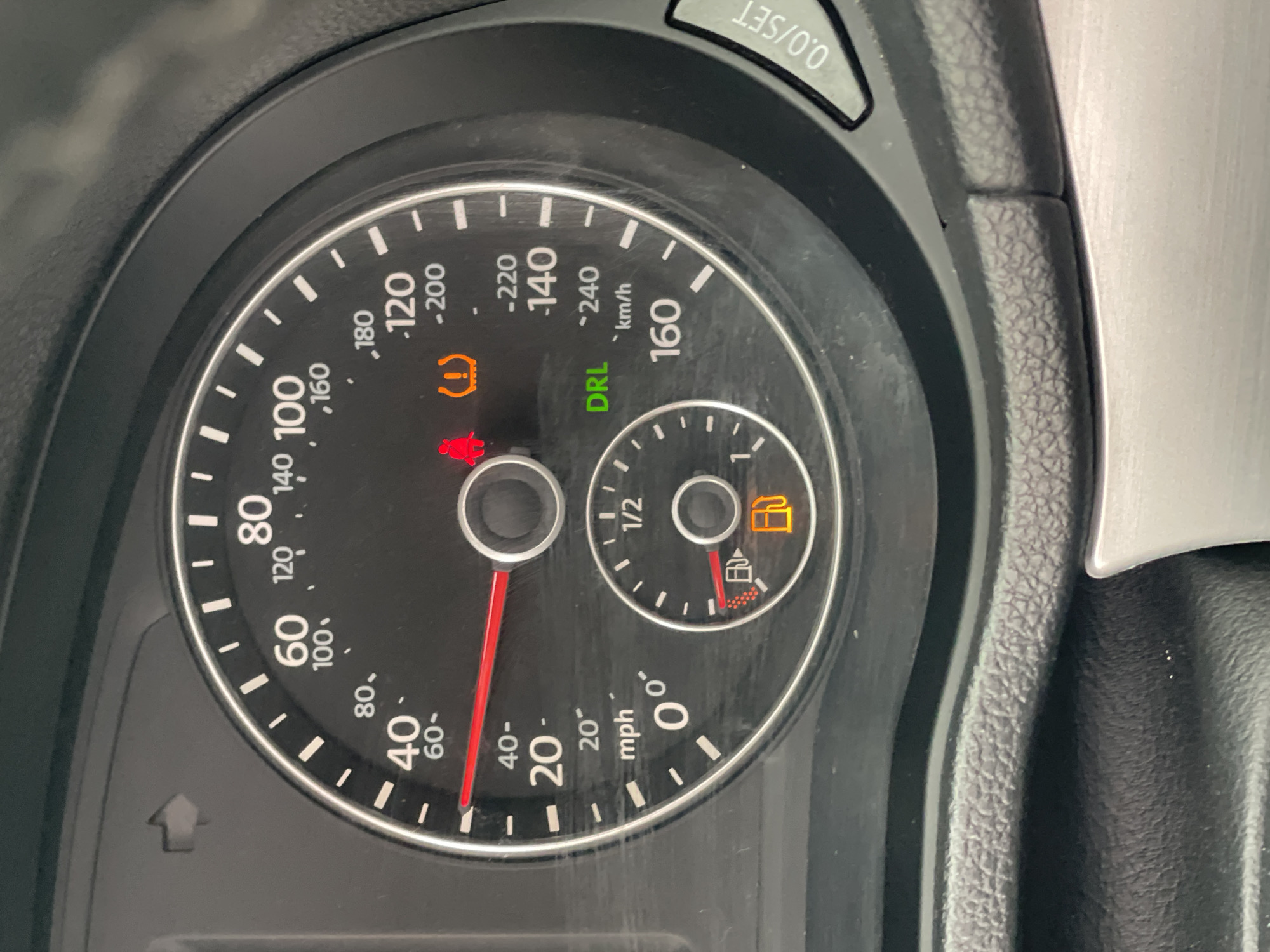 The image displays a close-up view of a car's instrument cluster, likely taken inside a vehicle in Kyiv, Ukraine. The speedometer shows a speed reading of approximately 25 mph, indicated by the red needle pointing between the 20 and 40 mph markers. The fuel gauge shows the tank is nearly full. Several warning lights are illuminated: a red tire pressure warning light with an exclamation mark inside a tire, and a green "DRL" indicator, likely signifying daytime running lights are active. A fuel pump icon is also illuminated in yellow, indicating low fuel. The instrument cluster is black with white markings and includes smaller gauges for fuel and possibly engine temperature. The surrounding dashboard is made of textured black plastic. The image is well-lit, with no visible people.