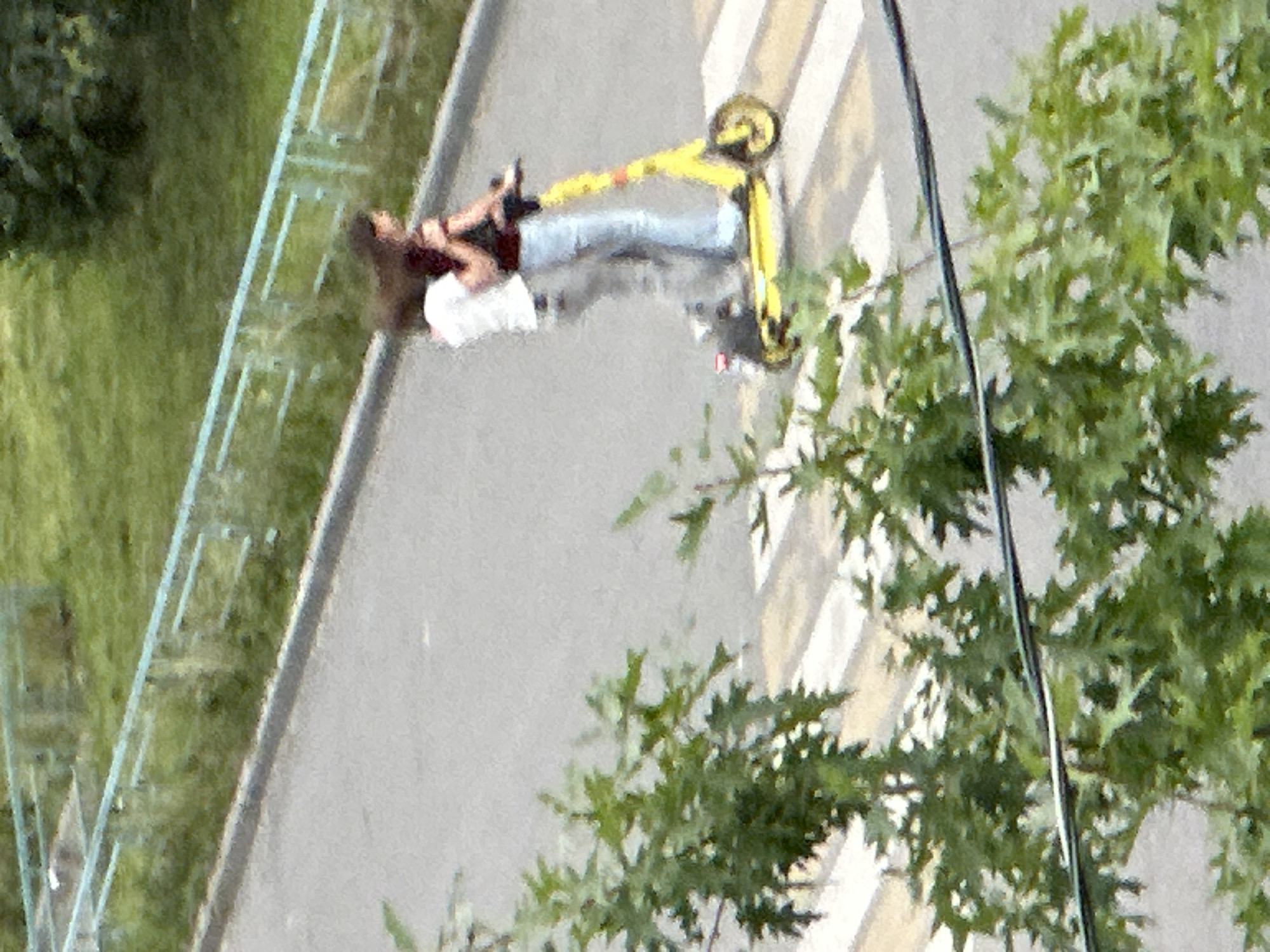 A person is lying on a zebra crossing in Podolsk, Russia. They are wearing a dark shirt and light-colored pants, with a white shirt underneath. Their body is oriented along the stripes of the crosswalk. Next to them, a yellow electric scooter is lying on its side, also across the zebra crossing. The person's left leg is bent at the knee, and their arms are partially obscured.
The scene appears to be outdoors, with a sidewalk and a grassy area with a fence visible to the left. Trees with green leaves are present on the right side of the image, casting shadows on the ground. The lighting suggests it could be daytime, possibly late morning or afternoon, given the brightness. The overall impression is of an unexpected situation, with the person and scooter obstructing the pedestrian crossing. There is no visible text in the image.