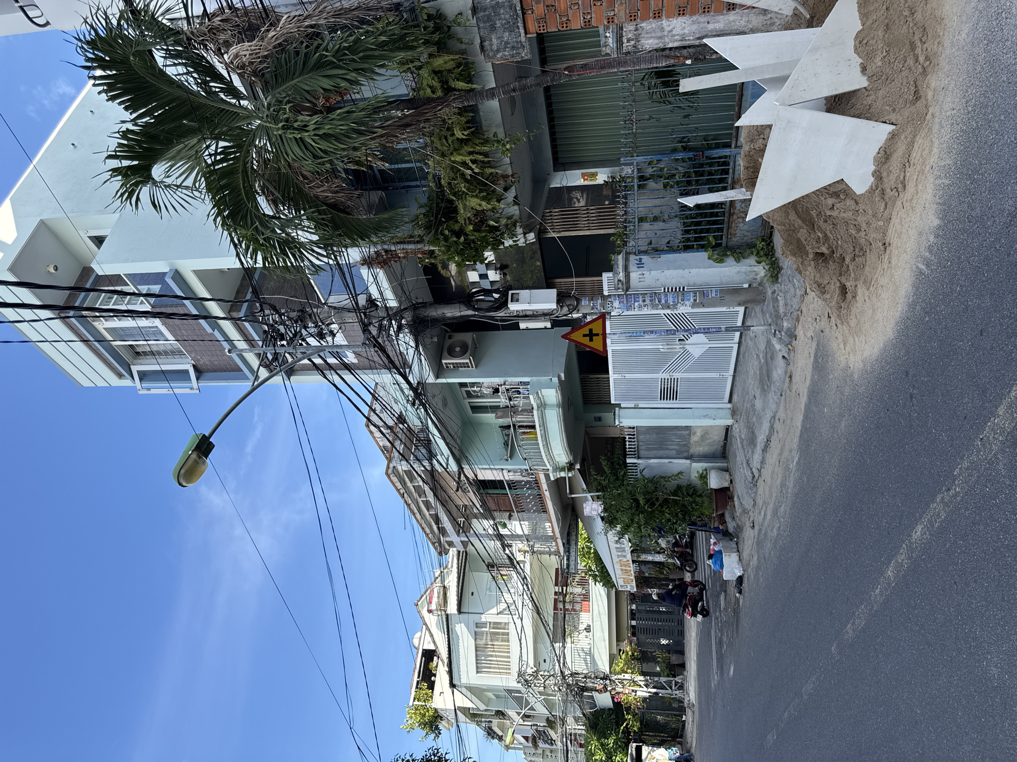 The image depicts a street scene in Nha Trang, Vietnam. In the foreground, a paved road with a white line is visible. To the right of the road, a pile of sand with several white, angular slabs is present. Adjacent to the sandpile, a fence made of dark metal bars encloses a property. Behind the fence, a building with a light blue facade and a green metal garage door is situated. A palm tree with green fronds is prominently positioned in front of this building.
Further down the street, several multi-story buildings, predominantly in pastel colors, line the left side. These buildings are adorned with balconies and numerous electrical wires are strung between them, creating a complex network overhead. A street lamp with a green fixture is visible in the upper left quadrant of the image. On the street, a person is riding a red motorbike, and another motorbike is parked. Potted plants and shrubs are placed along the sidewalk and in front of some of the buildings. The sky is clear and blue, indicating daytime. The scene suggests a residential or commercial area within the city.