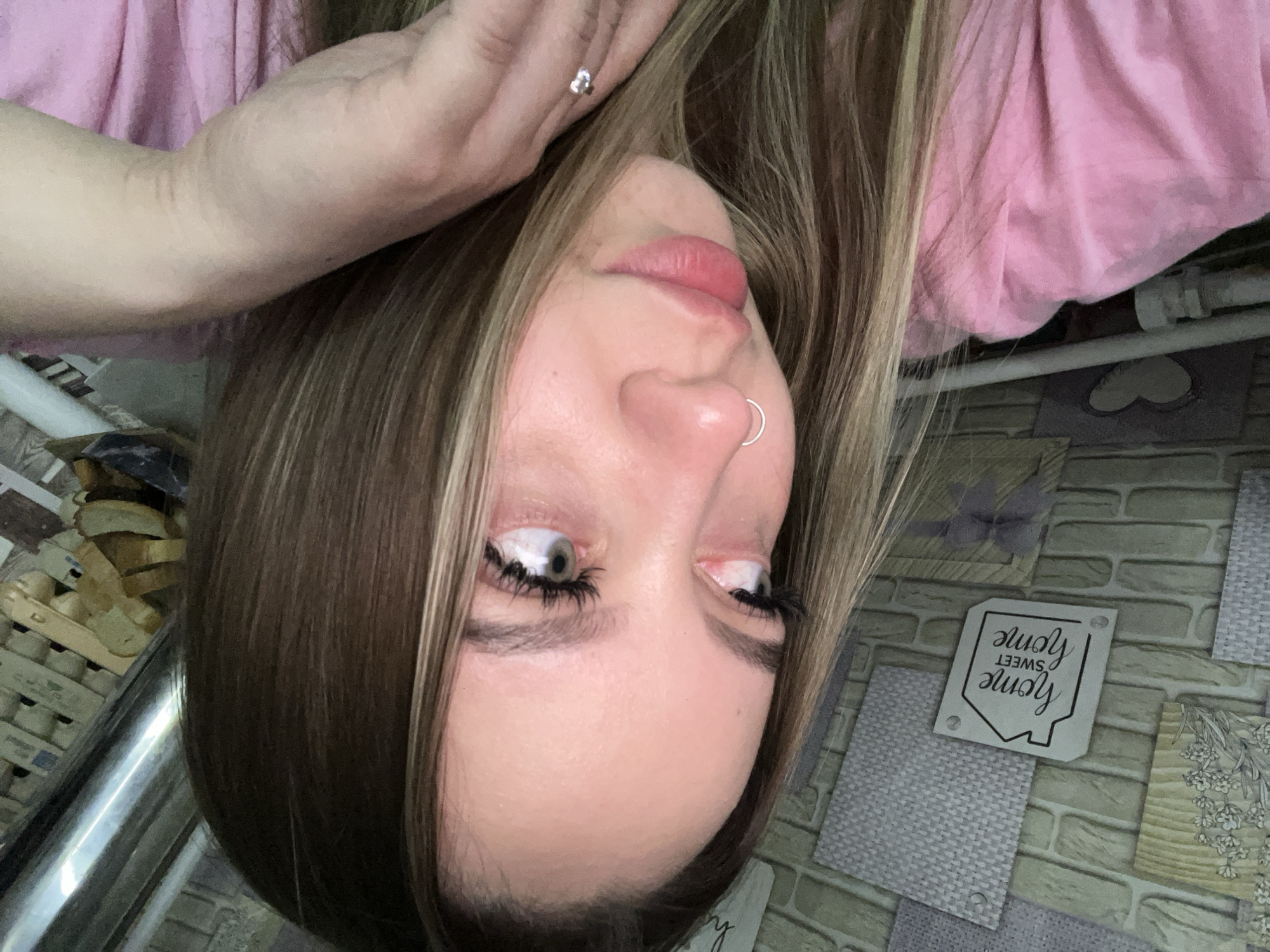 A woman with long brown hair and a nose ring is shown from an upside-down perspective, wearing a pink shirt. Her right hand is raised to her forehead and a ring is visible on one of her fingers. The background includes a metal pole, a shelf with stacked items, and a patterned wall with a "Home Sweet Home" decorative sign. The location appears to be indoors. The city and specific location within Port-Katon, Russia cannot be determined from the image.