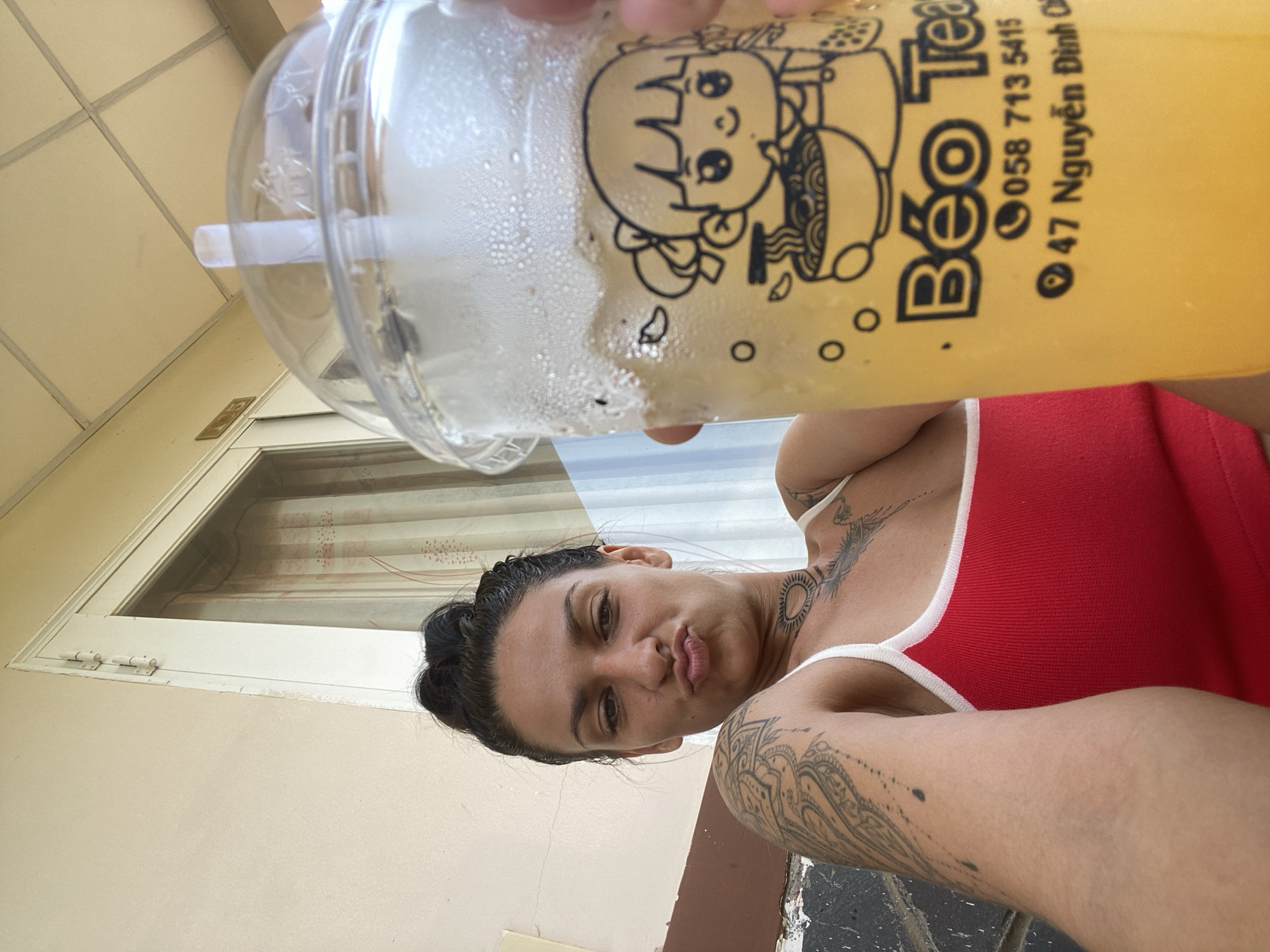 A woman with dark hair pulled into a bun, wearing a red tank top, makes a pouting face as she holds up a plastic cup of a yellow beverage. The cup has a cartoon image of a girl eating noodles and text that reads "Béo Tea" with a phone number and address. She has tattoos on her arms, chest, and neck. The background shows a light-colored wall, a doorway with a white frame, and a glimpse of a patterned curtain. The scene appears to be outdoors, possibly on a porch or balcony, with a light-colored ceiling overhead. The overall lighting suggests it is daytime.