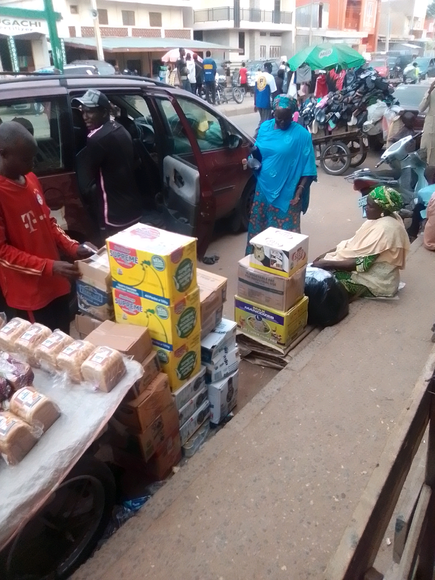 The image depicts a street scene in Arawa, Nigeria, likely a market area. In the foreground, a stall is laden with various food items, including loaves of bread wrapped in plastic and stacks of boxed goods. Behind the stall, a dark-colored van is partially visible with its door open. A young boy in a red t-shirt stands next to the van. Further back, a woman in a blue headscarf and patterned dress is standing near a cart piled high with what appear to be shoes, covered by a green umbrella. Several other individuals are present in the background, some seemingly engaged in commerce or passing by. The environment suggests a bustling urban setting with buildings and other vehicles in the distance.