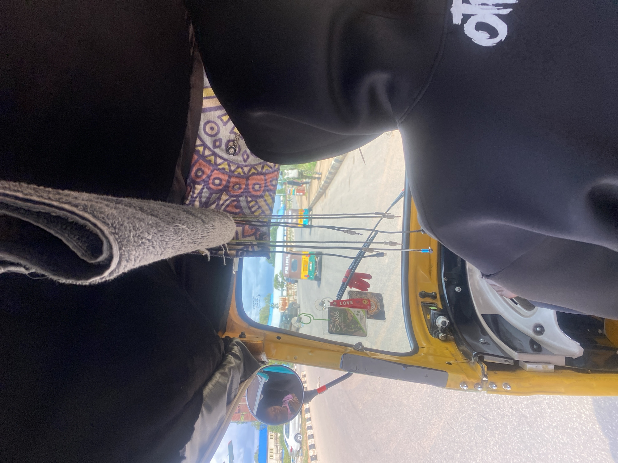 The image captures the interior of a yellow auto-rickshaw, likely in Barakin Lerawi, Nigeria. The perspective is from the passenger seat looking forward through the windshield. In the foreground, the back of a person wearing a black hooded sweatshirt with a white graphic design on the chest is visible. Dangling from the rearview mirror are several keychains, including one that says "Love" with a smiley face emoji. A circular side mirror is attached to the left side of the windshield, reflecting a person in a pink shirt.
Outside the vehicle, the road is paved, and there are other vehicles visible, including a white car on the left and another auto-rickshaw in the distance. Buildings and trees can be seen along the roadside. The sky is bright, suggesting daytime. The overall scene is typical of transportation in an urban or semi-urban area in Nigeria.