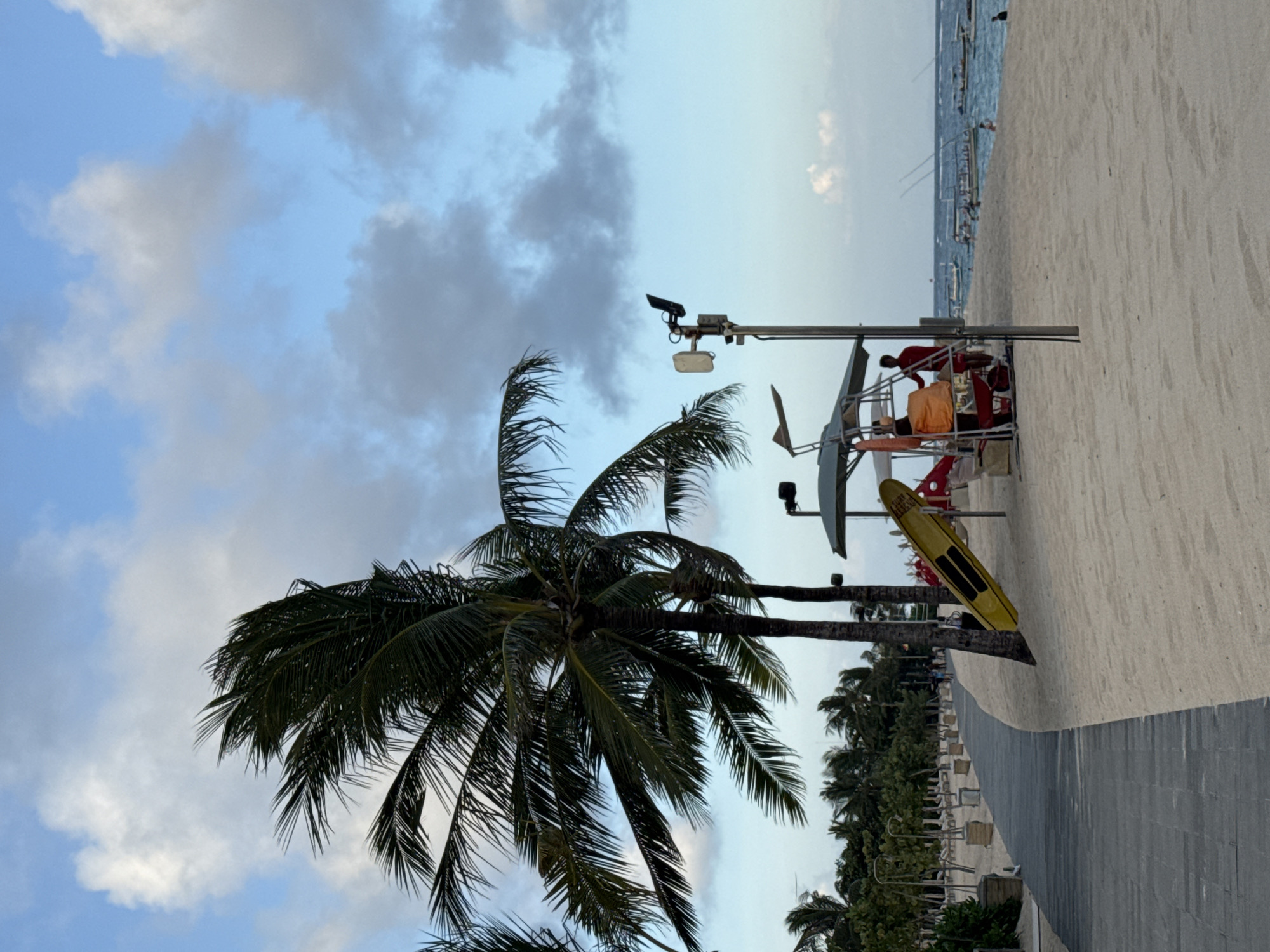 The image depicts a beach scene in Kuta Selatan, Indonesia, featuring a palm tree in the foreground against a partly cloudy sky. Adjacent to the tree, a lifeguard stand is visible, equipped with rescue gear, including a yellow surfboard. The stand appears to be occupied by at least one lifeguard, indicated by an individual in orange attire. A boardwalk runs parallel to the beach, bordered by rows of beach chairs and additional vegetation. The sandy beach extends towards the ocean.
