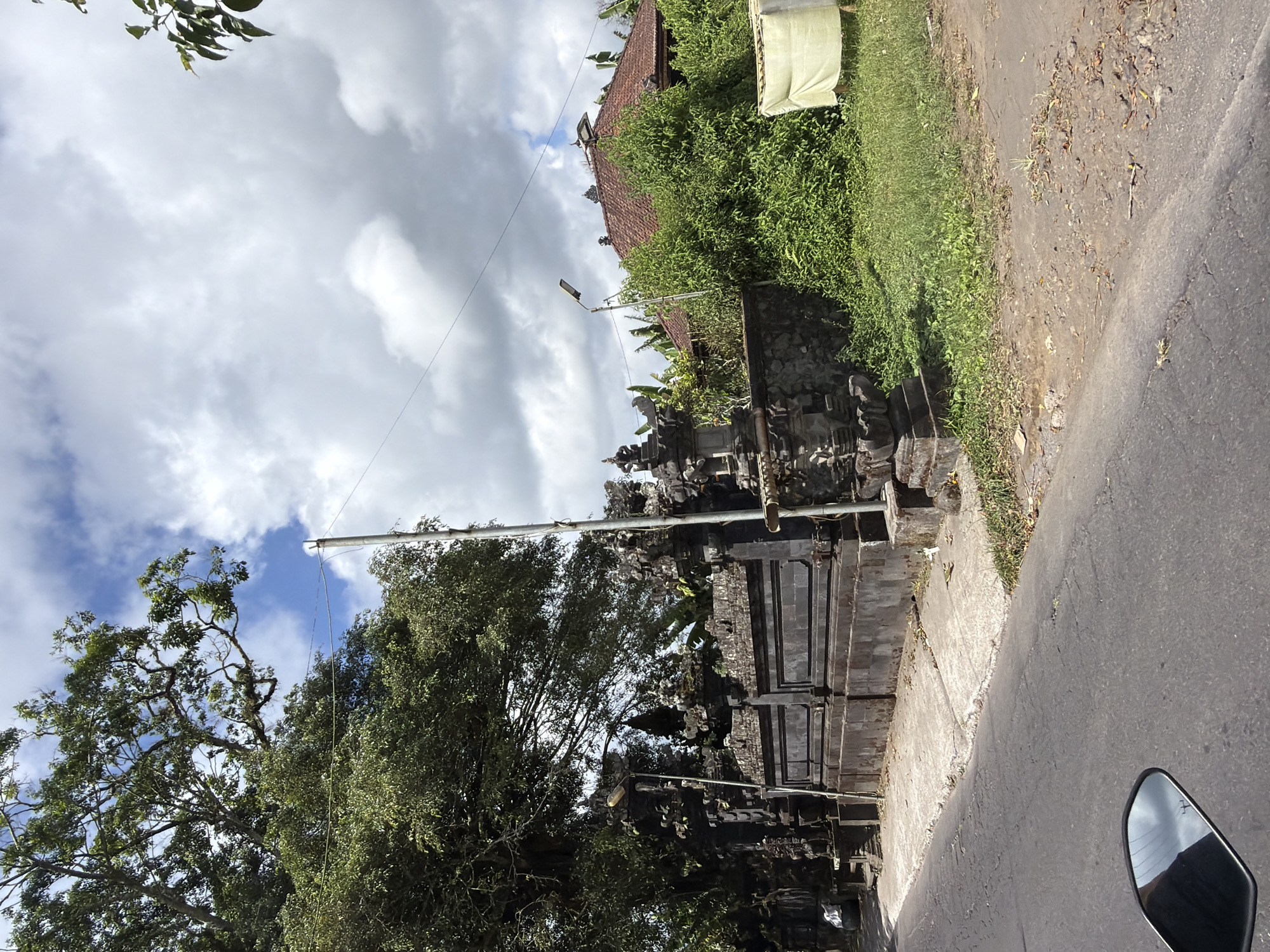 The image captures a roadside view in Kintamani, Indonesia, featuring a traditional Balinese temple gate and a residential building. The temple gate, constructed from dark stone with intricate carvings, is a prominent element on the left side of the frame. Adjacent to the gate, a large tree with lush green foliage extends its branches towards the sky. To the right of the temple, a house with a red tiled roof is partially obscured by dense green vegetation. A pale yellow tarp is visible in the far right of the frame, possibly covering something. The scene is set against a backdrop of a cloudy sky. The foreground shows a paved road with a motorcycle's side mirror visible in the lower left corner, reflecting the sky. The environment appears to be a mix of natural elements and cultural architecture, characteristic of rural Indonesian landscapes. There are no people visible in the image.