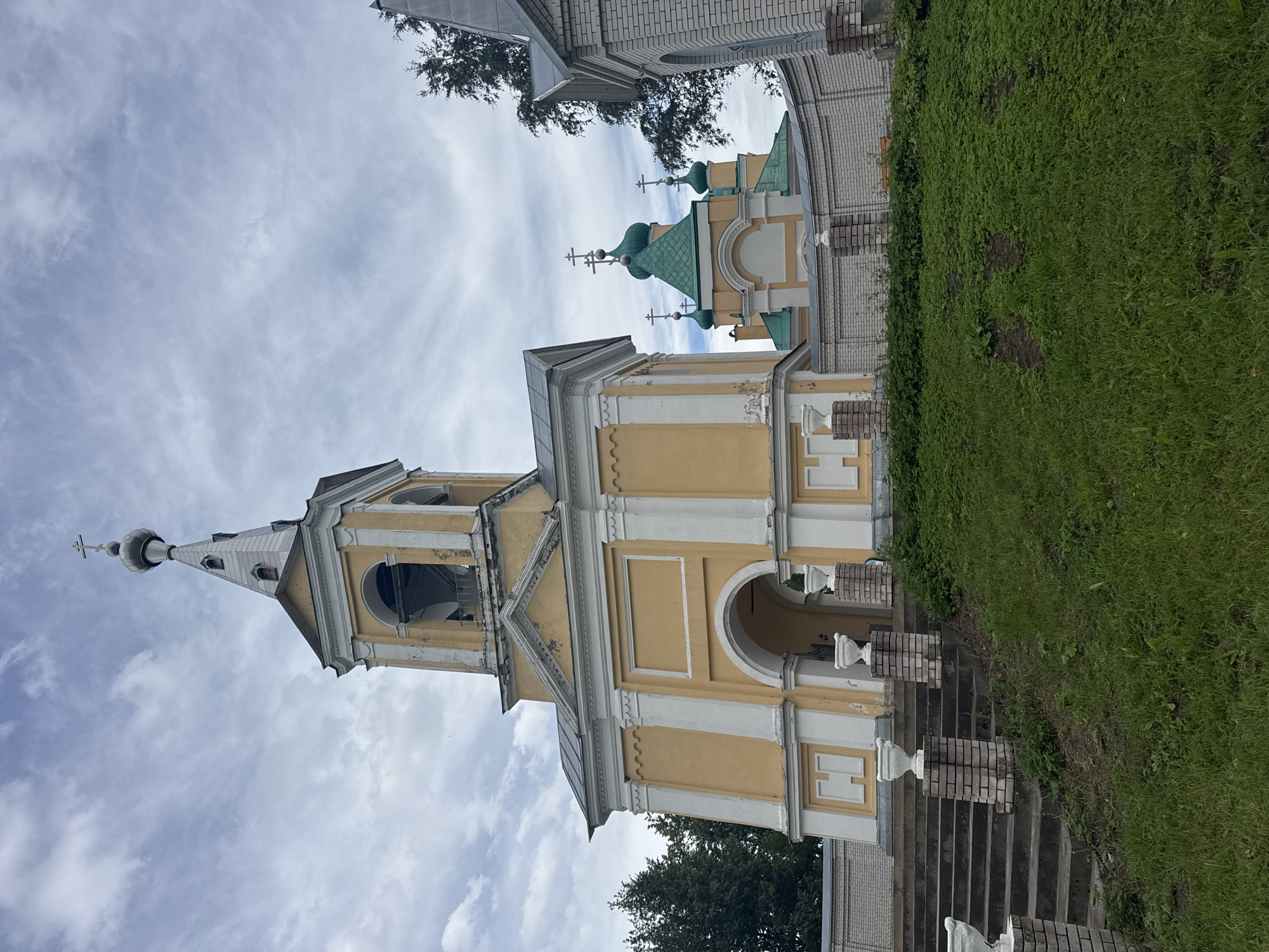 The image shows a view of a yellow bell tower and an Orthodox church with green domes in the city of Рассвет, Russia. The bell tower, decorated with white columns and arches, is prominently featured on the left side of the image. Behind it, a portion of the church, with its distinctive green domes topped with crosses, is visible. A white brick wall with decorative pillars encloses the church grounds. The foreground consists of a grassy slope leading up to a set of stone stairs that ascend towards the bell tower. The sky is overcast with scattered clouds, and trees can be seen in the background, adding to the serene atmosphere of the religious site. There are no people or animals visible in the scene.