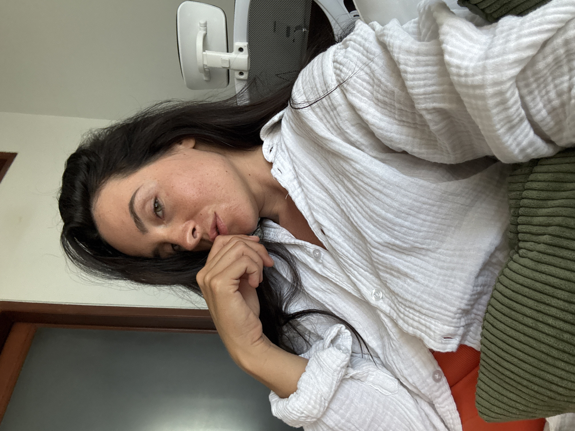 A medium shot captures a woman with dark hair and light skin from the chest up, seated indoors in a room with light walls. She is wearing a loosely buttoned, white crinkled fabric shirt with puffed sleeves. Her left hand is raised to her mouth, with her fingers resting on her lower lip and chin, and her gaze is directed slightly upward and to the right.
The background shows a portion of a wooden door frame and a white office chair with a mesh back. A green corduroy pillow rests in the foreground, partially obscuring an orange surface. The scene suggests a casual, indoor setting. The location is specified as North Kuta, Indonesia.
