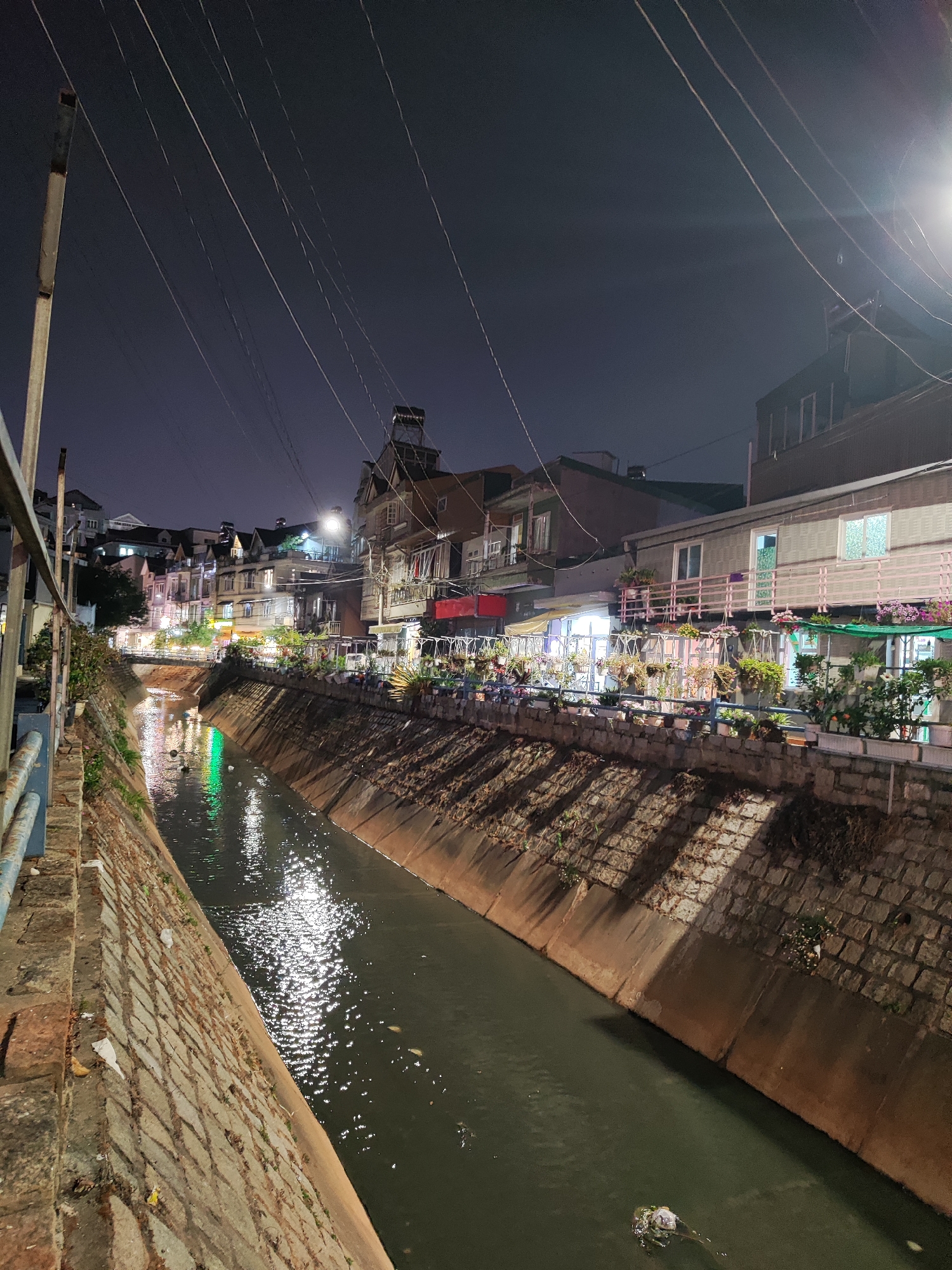 The photograph shows a canal in Cam Ly - Đà Lạt, Vietnam at night. The canal is lined with stone embankments and the water appears dark, reflecting light from street lamps and buildings. The banks on either side of the canal are lined with buildings, mostly residential. The buildings have flower boxes and other plants. Overhead, a web of electrical wires stretches across the scene. The lighting is a mix of bright artificial light from the buildings and street lamps, and the overall impression is that of a quiet urban scene. Some garbage is visible in the water.