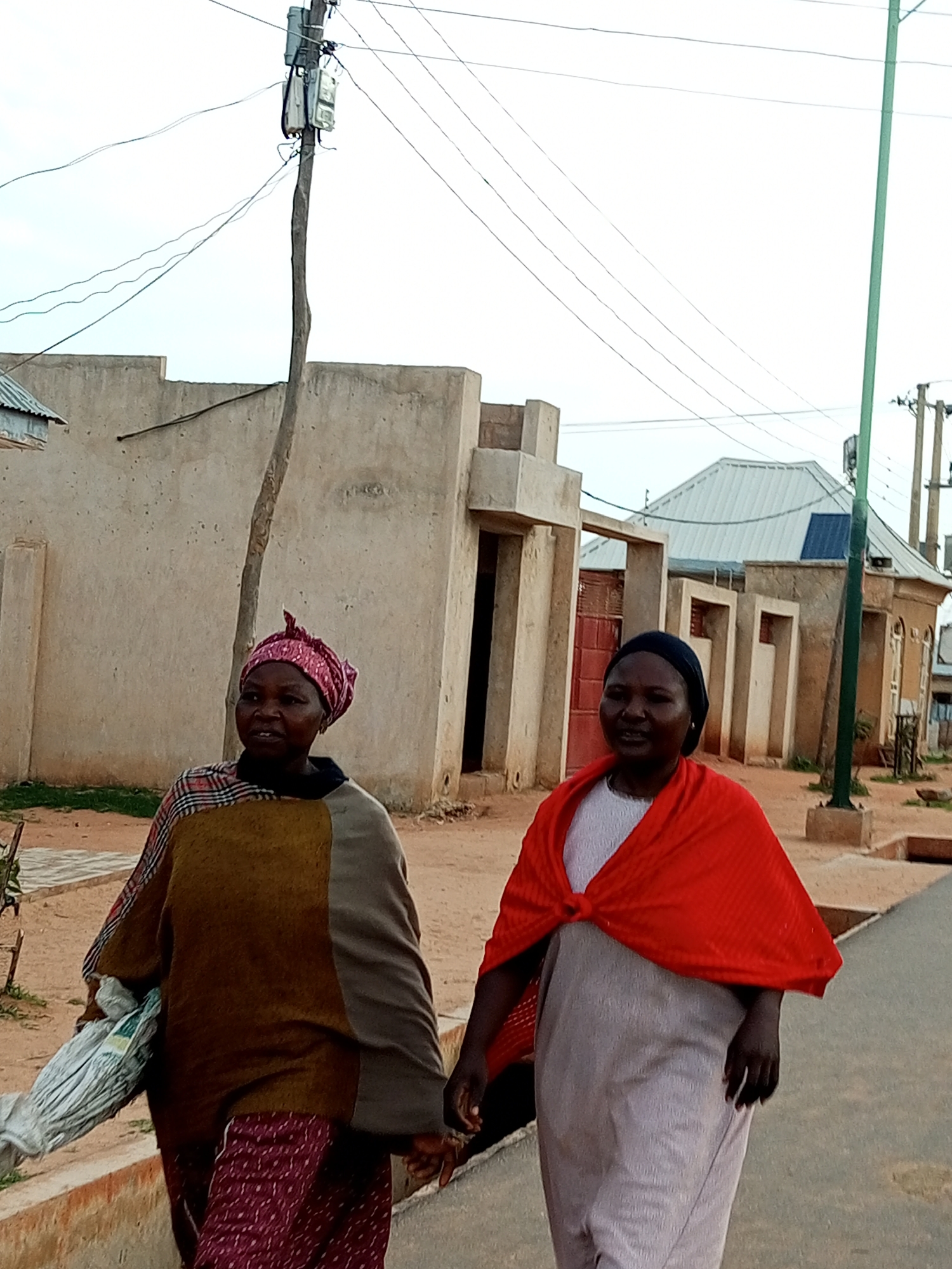 Two women walk side-by-side outdoors in Gombe, Nigeria. The woman on the left wears a pink headscarf and a patterned shawl over a maroon skirt. She carries a bundled item in her left arm. The woman on the right wears a black head covering and a bright red shawl over a light-colored dress. They appear to be walking on a paved path next to a dirt area. In the background, there are several light-colored buildings with simple architectural designs, and utility poles with wires crisscross the sky. The sky is overcast.