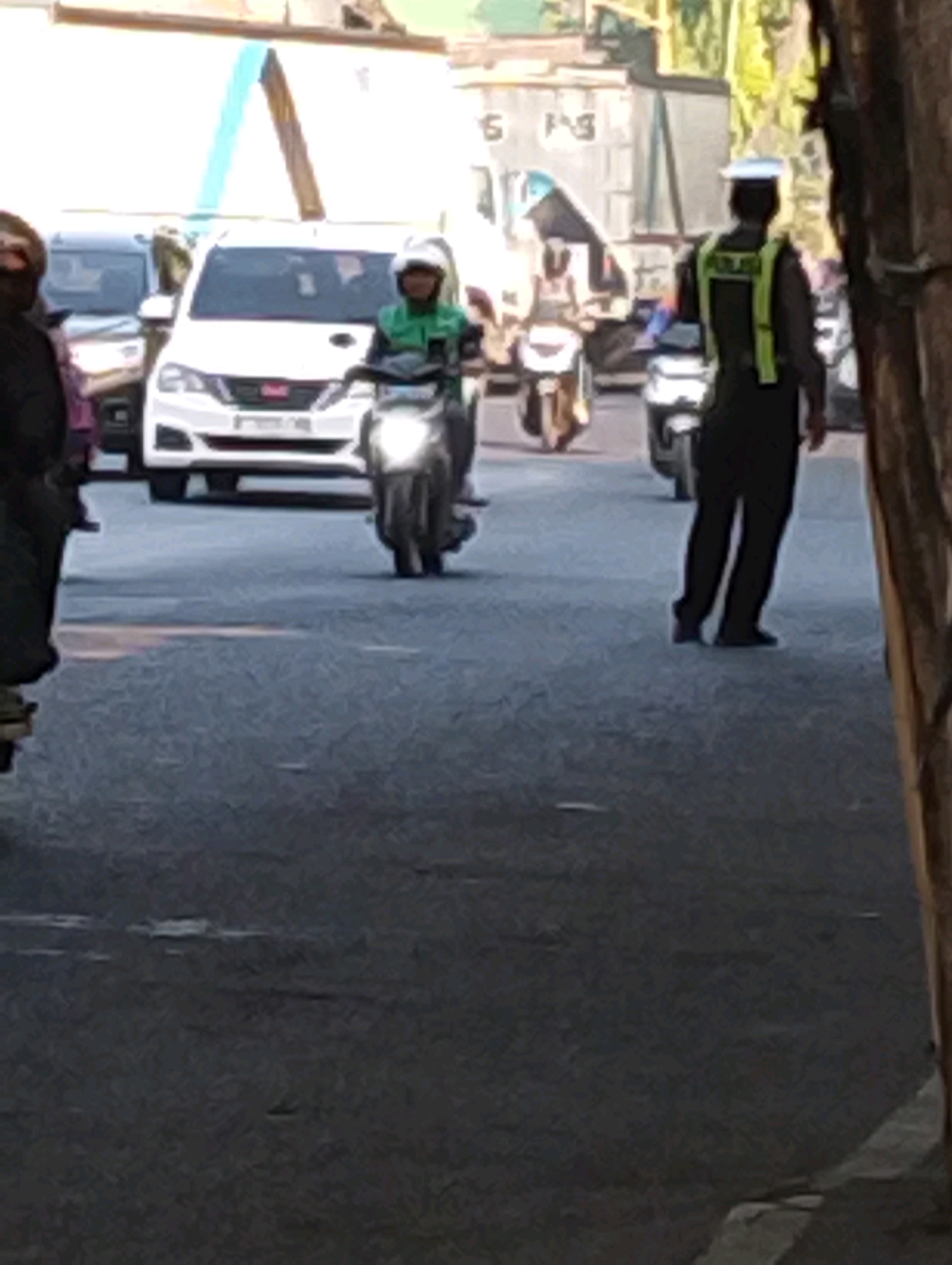 In Bangil, Indonesia, on an asphalt road under daylight, a traffic officer wearing a dark uniform with a bright yellow reflective vest and a white cap stands facing away from the camera. In front of him, a white car is parked, and to the left of the car, a person on a scooter with a green jacket and helmet is moving forward. Other vehicles and pedestrians are visible in the background, suggesting a busy street scene.