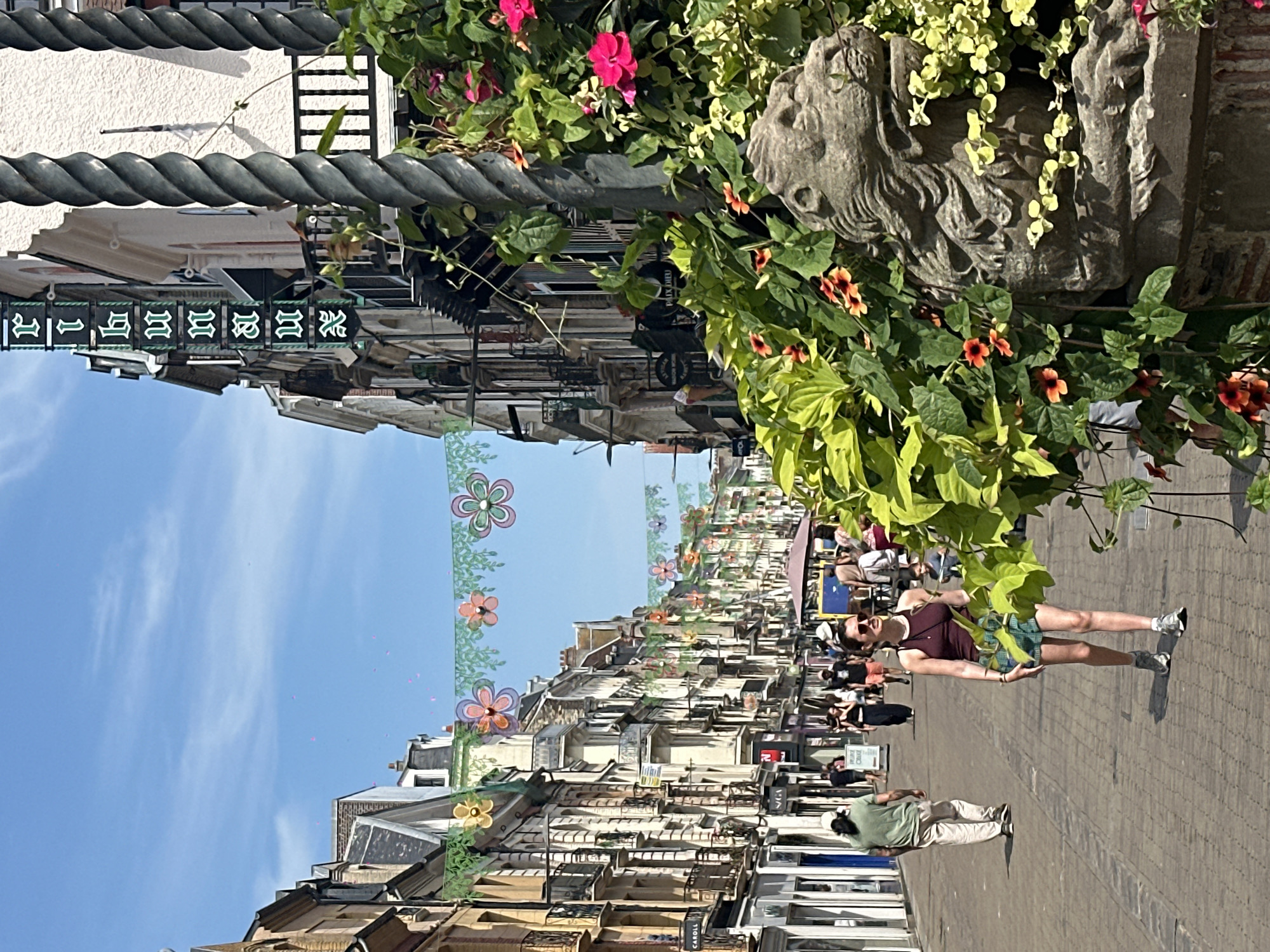The image depicts a sunny day on a pedestrian street in Dieppe, France, likely Rue Jacques-Émile Blanche or a similar commercial street. Buildings with traditional French architecture line both sides of the narrow street, featuring balconies and ornate facades. Decorative flower garlands are strung across the street overhead, with colorful fabric flowers in shades of pink, orange, yellow, and green. In the foreground, a woman with sunglasses, wearing a dark tank top and plaid shorts, walks towards the viewer. To her left, a man in a light green shirt and beige pants walks away from the viewer. Other pedestrians are visible further down the street, engaged in shopping or strolling. The street is paved with cobblestones. To the right of the frame, a decorative stone lion sculpture is partially obscured by lush green foliage and vibrant pink and orange flowers. A sign with green lettering on a dark background is visible on one of the buildings.