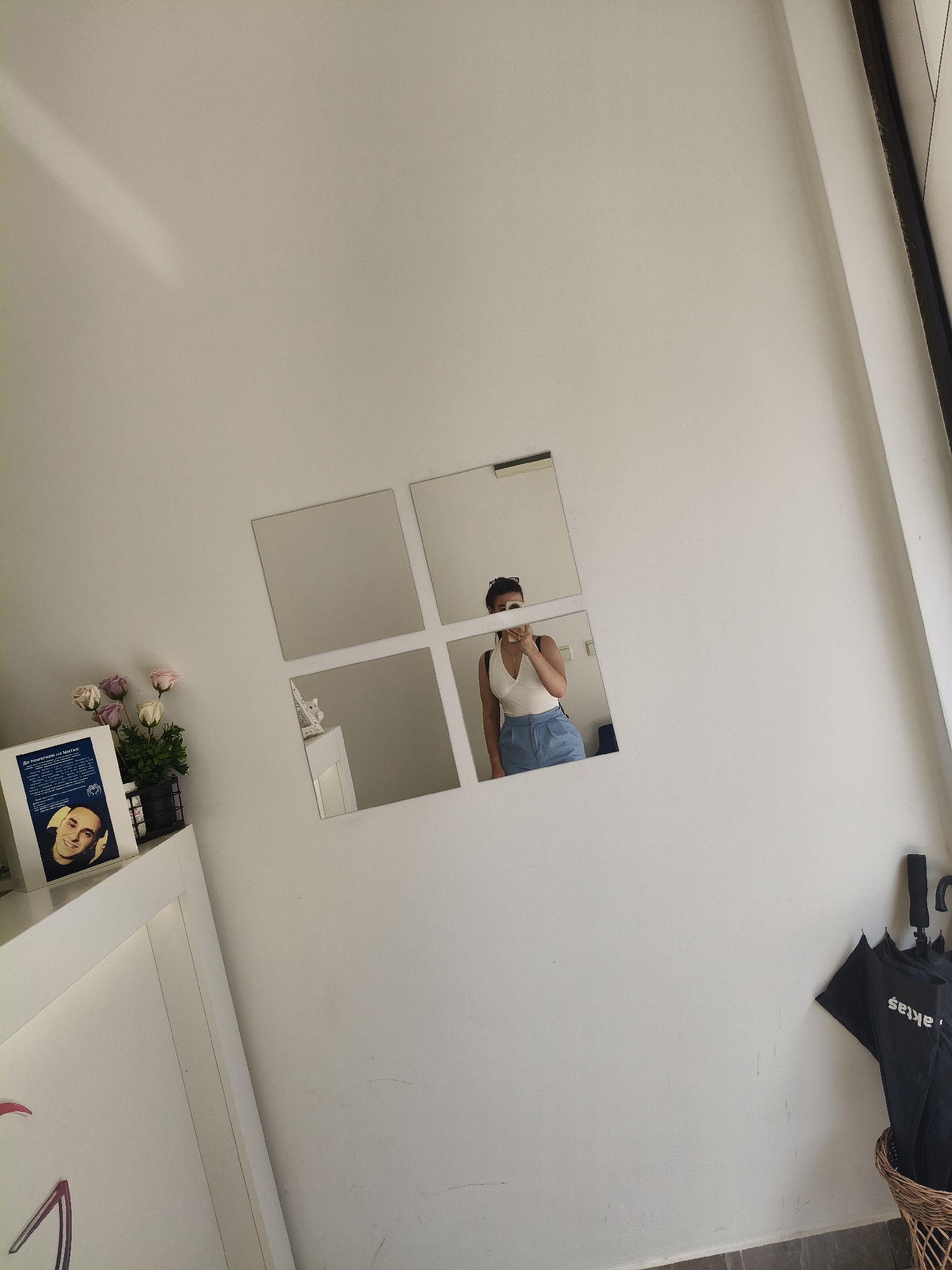 The image depicts an interior scene with a white wall as the primary background. Several square mirrors are affixed to the wall, arranged in a grid-like pattern that resembles the Windows logo. In one of the mirrors, a person is visible, taking a selfie. This individual is wearing a white halter top and blue shorts. To the left of the mirrors, a shelf holds a small potted plant with pink and white roses. Beside the plant is a framed document or picture featuring a portrait of a man. Further left, a white structure with a soft glow suggests a piece of furniture or architectural element. On the right side of the image, a wicker basket contains several black umbrellas, one of which has the word "aktas" printed on it. The overall impression is of a modern, minimalist interior space. The location is not specified in the image.