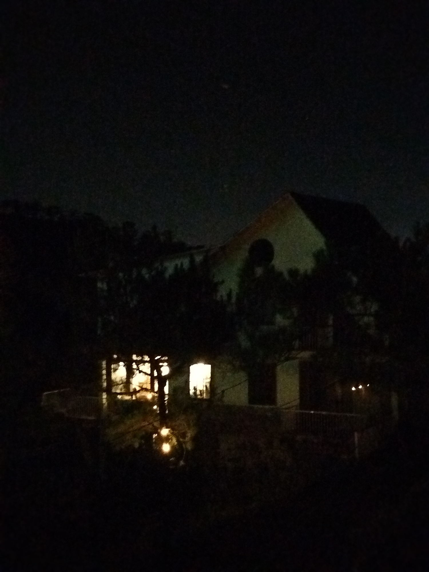 The image depicts a dimly lit, possibly nighttime scene. A building or structure is partially visible amidst dark foliage. The building appears to have multiple windows illuminated from within. There are decorative lighting elements in the foreground. The scene appears to be residential or recreational. There is no evidence of people, specific objects beyond those described or actions. The environment is dark and obscured. There is no indication of a location in Lạc Dương, Vietnam.