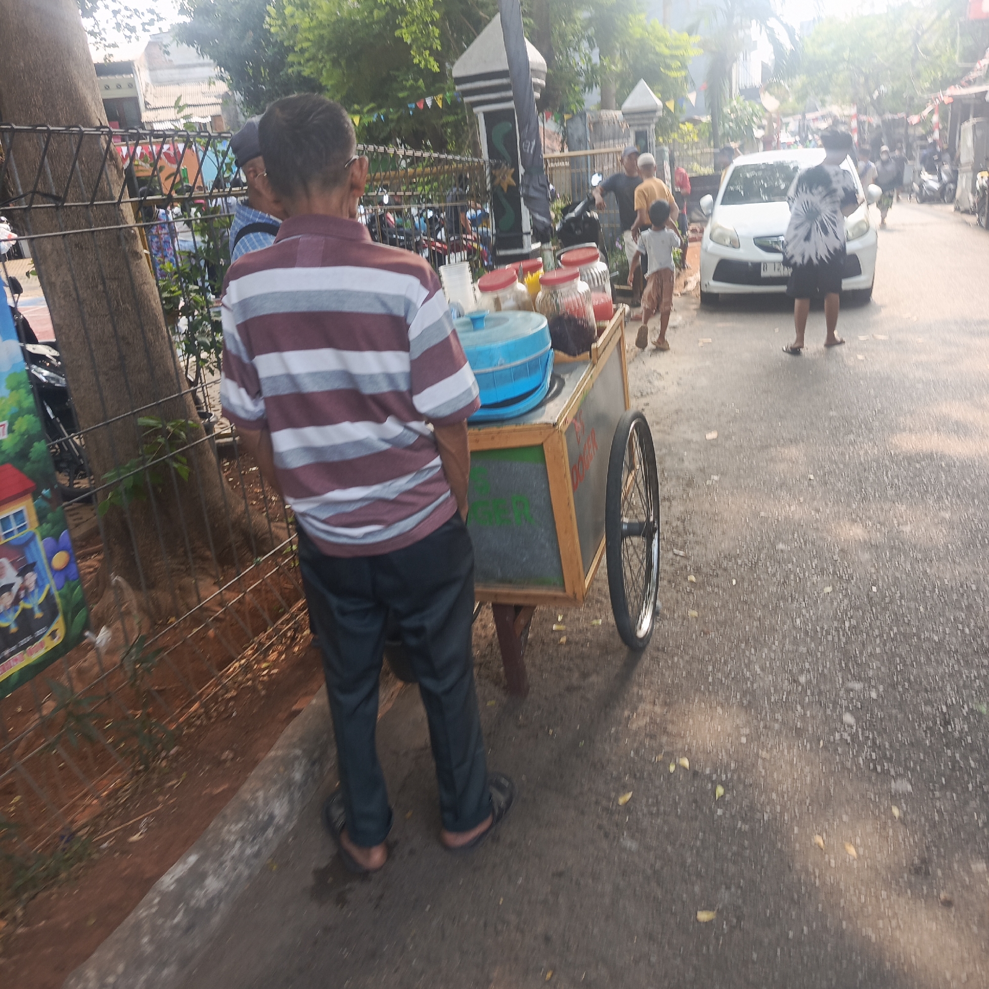 A man in a striped shirt and dark pants stands next to a food cart on a street in South Jakarta, Indonesia. The cart has a blue cooler and glass jars filled with colorful drinks. A white car is parked in the background, and several people are visible on the street. The scene appears to be during the daytime, with sunlight illuminating the area. The environment suggests a typical urban street scene with buildings and trees lining the road.