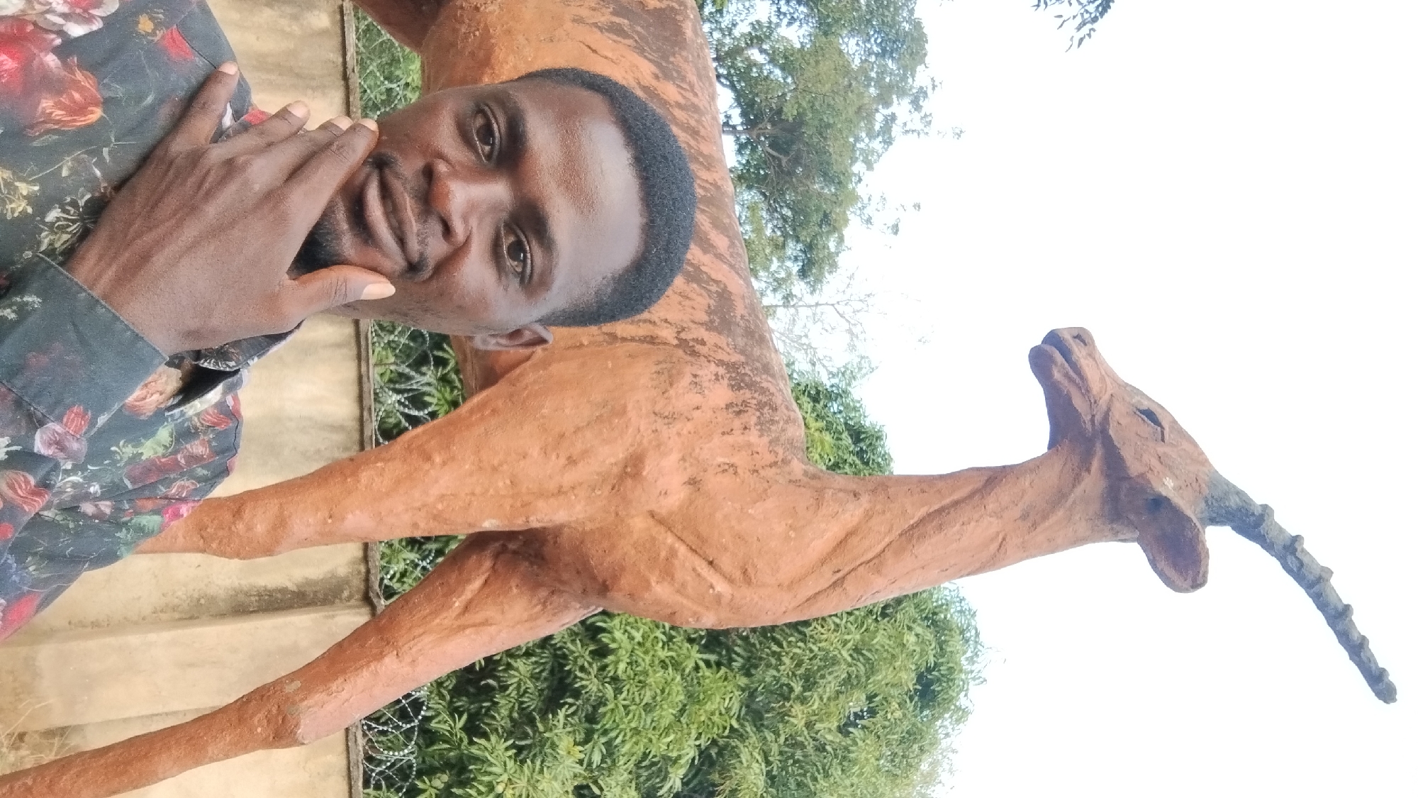 A man poses in a selfie with a life-size antelope statue. The man is dark-skinned with short black hair and a trimmed beard. He wears a floral patterned shirt and his hand is near his face. The statue is brown and weathered, with a long neck and a single horn. In the background, there is green foliage, trees, and a bright sky. Based on general information available, this scene is consistent with the city of Jos, Nigeria, known for its parks and sculptures.