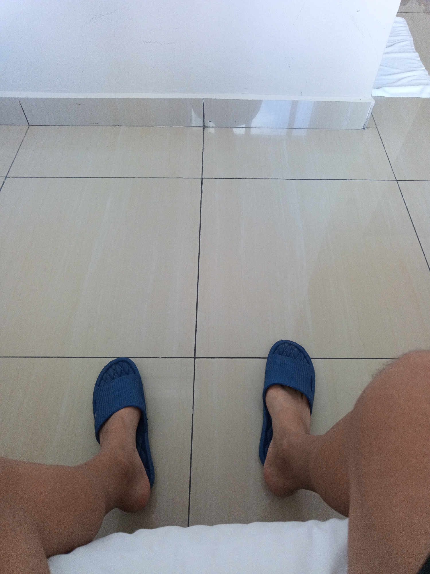 The image shows a first-person perspective of a person's legs and feet wearing blue slippers, resting on a tiled floor. The floor tiles are beige and arranged in a grid pattern. A white wall with a baseboard runs along the top of the image. Part of a white bedsheet or towel is visible in the foreground. The scene appears to be indoors, possibly a hotel room, in Kuala Lumpur, Malaysia.