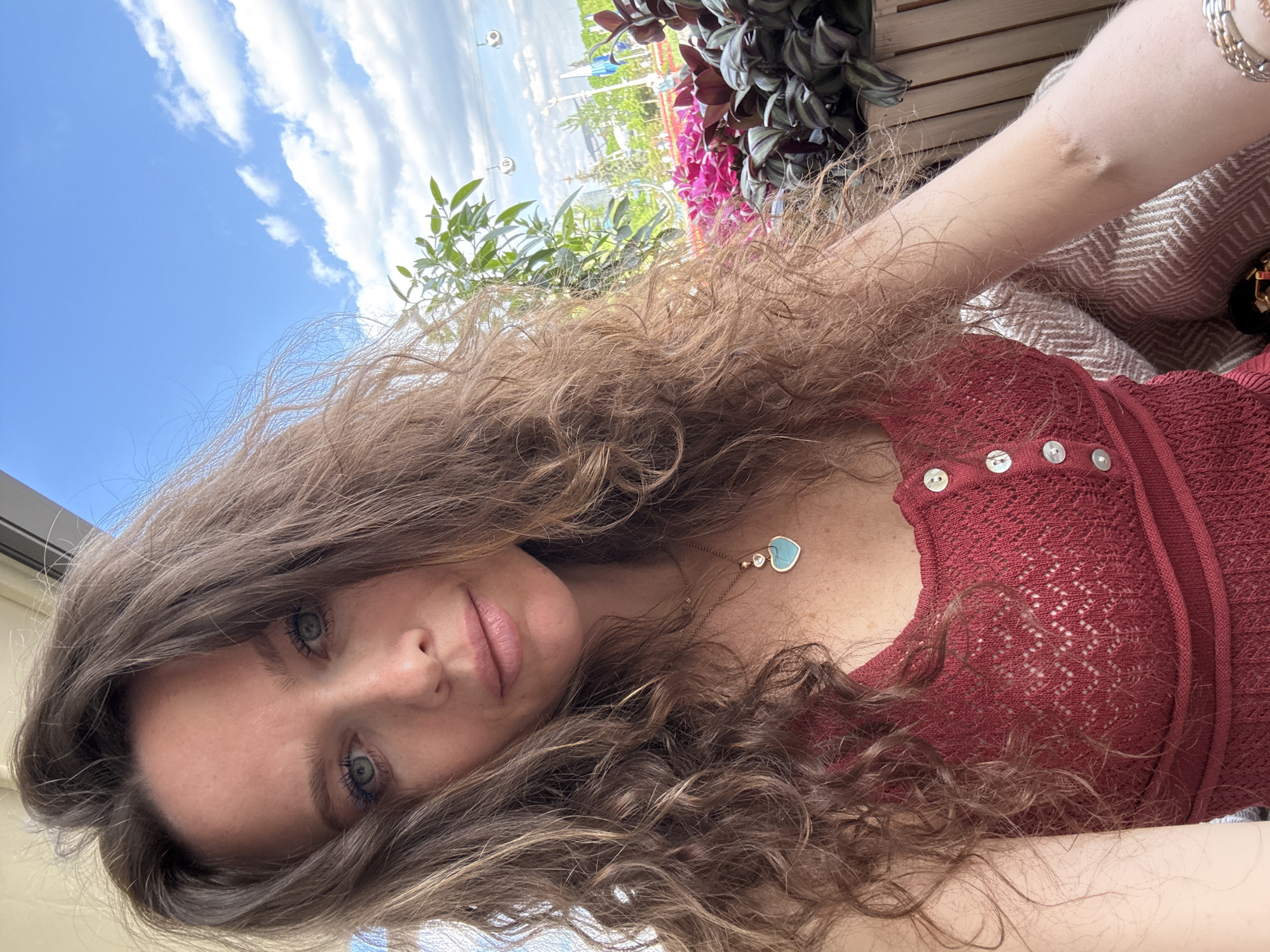 A woman with long, wavy brown hair, blue eyes, and light skin is depicted in a close-up, slightly angled selfie. She is wearing a red, knitted top with mother-of-pearl buttons. A necklace with a heart-shaped turquoise pendant is visible. The background shows a partly cloudy sky and greenery, including potted plants with pink and purple flowers. In the distance, a cityscape is visible, partially obscured by the greenery. Based on the architectural style and distinct structures in the background, the location appears to be Moscow, Russia.