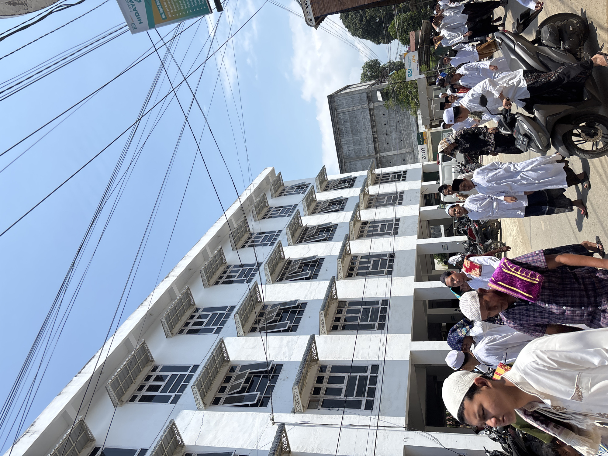 The image captures a bustling street scene in Mideuenjok, Indonesia, on a bright, sunny day, viewed from a low angle looking upwards. The clear blue sky is crisscrossed by numerous black electrical wires.
On the left, a prominent multi-story white building features many rectangular windows, some of which are open, and ornate, patterned awnings or small balconies on each level. To the right, a line of men and boys, predominantly dressed in white Islamic garments such as kurtas/thawbs and kufis/pecis, are either walking or standing alongside parked motorcycles. Some individuals wear patterned sarongs or shirts. They appear to be engaged in a gathering or procession.
In the background, behind the white building, a concrete, possibly unfinished, structure is visible. Further right, along the street, other commercial buildings are present. Several motorcycles (scooters) are parked on the side of the road, with some of the men either standing next to them or seated on them.
Visible text includes "BSI SMART FOTOC FOTO HIDAYA" on a green sign that also lists services like "MELAYANI Transfer, Setor T, Bayar, Toker, Pulsa, Data, Pak, Tog." Other signs display "BSI ATM" and "atm." A motorcycle license plate reads "BL5055 ZBB."
