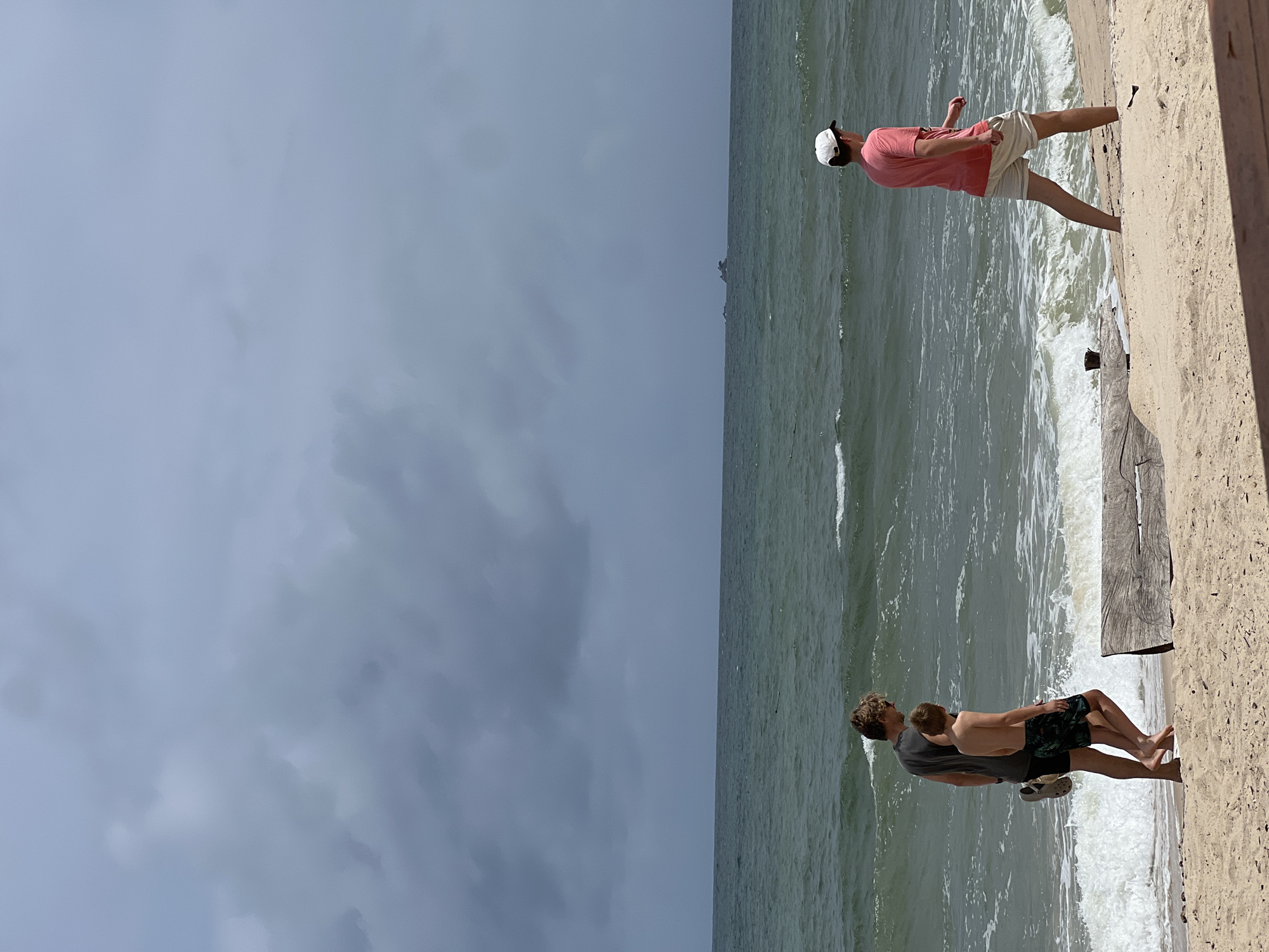 Three people are walking along a sandy beach on a cloudy day. The two in the foreground on the left are a taller adult male wearing a dark t-shirt and black shorts, holding a grey sandal in his left hand, and a younger male wearing a dark patterned swim trunk. They are walking side-by-side towards the right, with their backs to the camera. The third person, a young male wearing a pink t-shirt and light-colored shorts and a white baseball cap, is walking further ahead and to the right, also with his back to the camera. They are on a beach with sand in the foreground and a wooden log lying horizontally in the sand. The ocean is to the left, with choppy waves and a distant, dark, rocky island in the middle ground. The sky is filled with heavy, dark grey clouds.