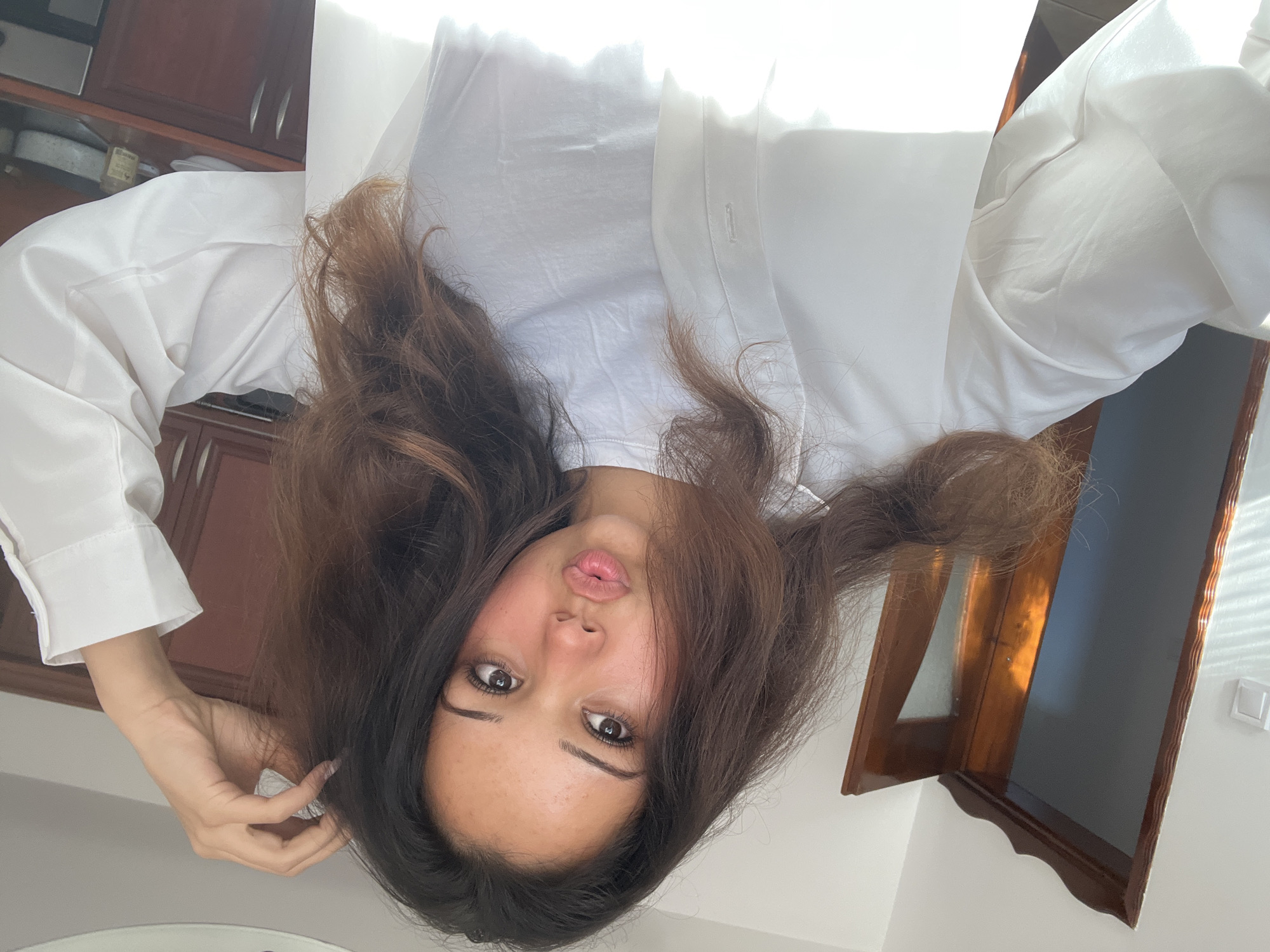 A young woman with long, dark, wavy hair is taking a selfie indoors, possibly in Zlatograd, Bulgaria. She is wearing a white collared shirt over a white t-shirt and puckers her lips for the camera. Her right hand is raised and touching her hair. The background shows a doorway with a dark wooden frame and a glass pane, kitchen cabinets, and natural light streaming in, creating bright highlights and shadows. A small white object is mounted on the wall to the left of the doorway.