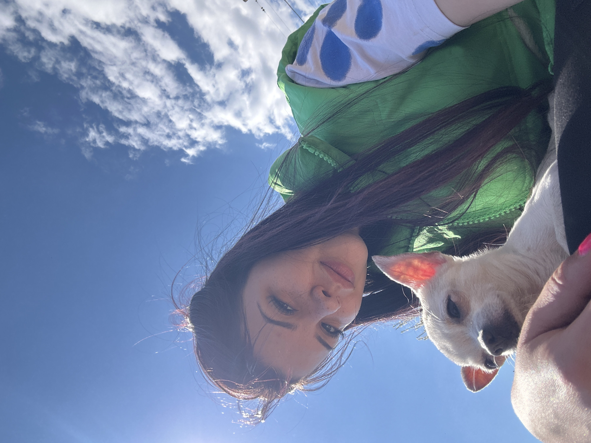 A low-angle photograph shows a woman holding a white dog against a backdrop of blue sky and scattered clouds. The woman has dark hair, fair skin with freckles, and is wearing a green jacket over a white shirt with blue polka dots. Part of her hand with pink nail polish is visible. The dog is small with short white fur and pink ears. The location is not discernible, and there is no evidence the photo was taken in Chubra, Bulgaria.