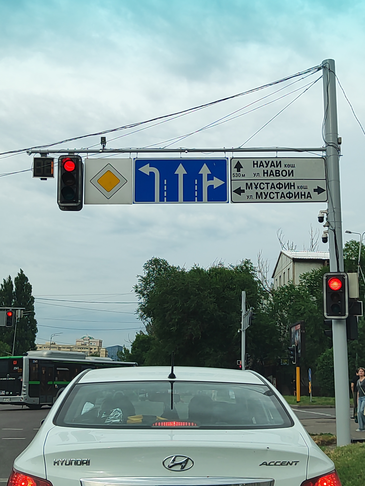 The image shows a street scene in Almaty, Kazakhstan, likely at an intersection. In the foreground, a white Hyundai Accent car is visible from the rear. Behind it, a bus with the number 2541 on its side is driving. Above the road, traffic lights are present, with a red light illuminated. There are also street signs indicating directions: one with arrows for turning left, going straight, and turning right, and another sign with text in Kazakh and Russian. The text on the sign indicates "NAYAI kosh" (likely a street name) 530 meters ahead, and "UL. NAVOI" (Navoi Street), and also "MUSTAFIN kosh" (Mustafin Street) and "UL. MUSTAFINA" (Mustafina Street) with directional arrows. Trees and buildings are visible in the background, under a cloudy sky. Two security cameras are mounted on a pole near the traffic lights. A person is walking on the sidewalk on the right side of the image.