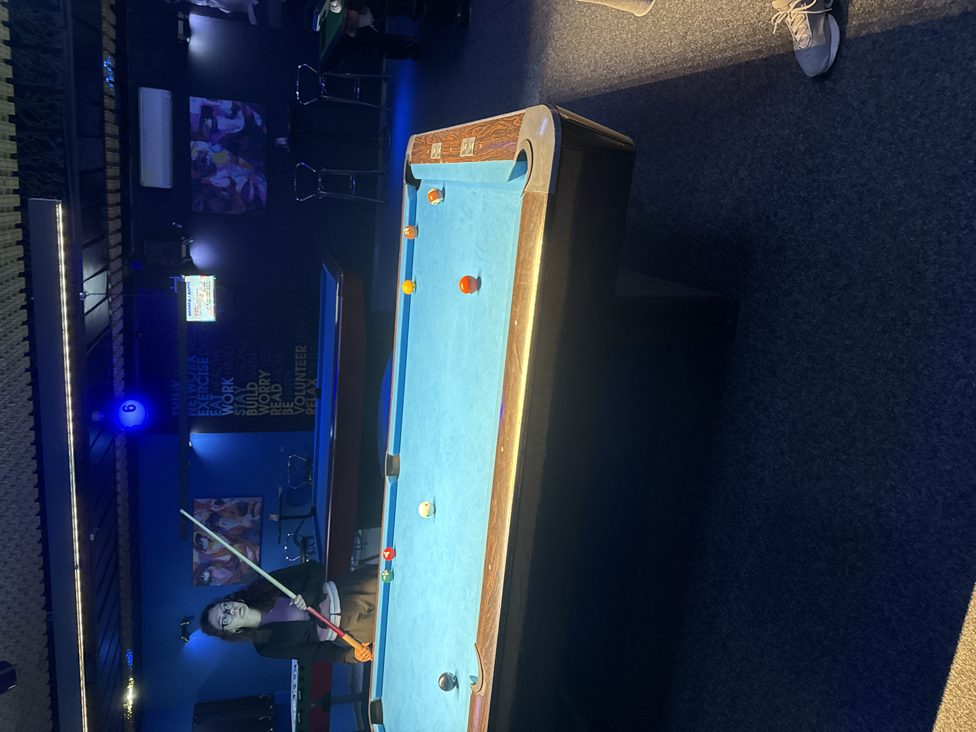 The image captures an indoor scene, likely a pool hall, with a prominent blue pool table in the foreground. A young woman with dark hair and glasses stands to the left of the table, holding a pool cue. She is dressed in a black jacket over a purple top and dark pants. Several billiard balls are scattered on the table, including a black ball, a solid red ball with the number 4, a yellow-green ball with the number 3, and a white cue ball. In the background, a wall is decorated with abstract art and text. A sign with the number 6 is illuminated overhead. The room appears to be dimly lit, with blue ambient lighting creating a mood. The floor is carpeted. No specific Yambol, Bulgaria landmarks are identifiable within the image.