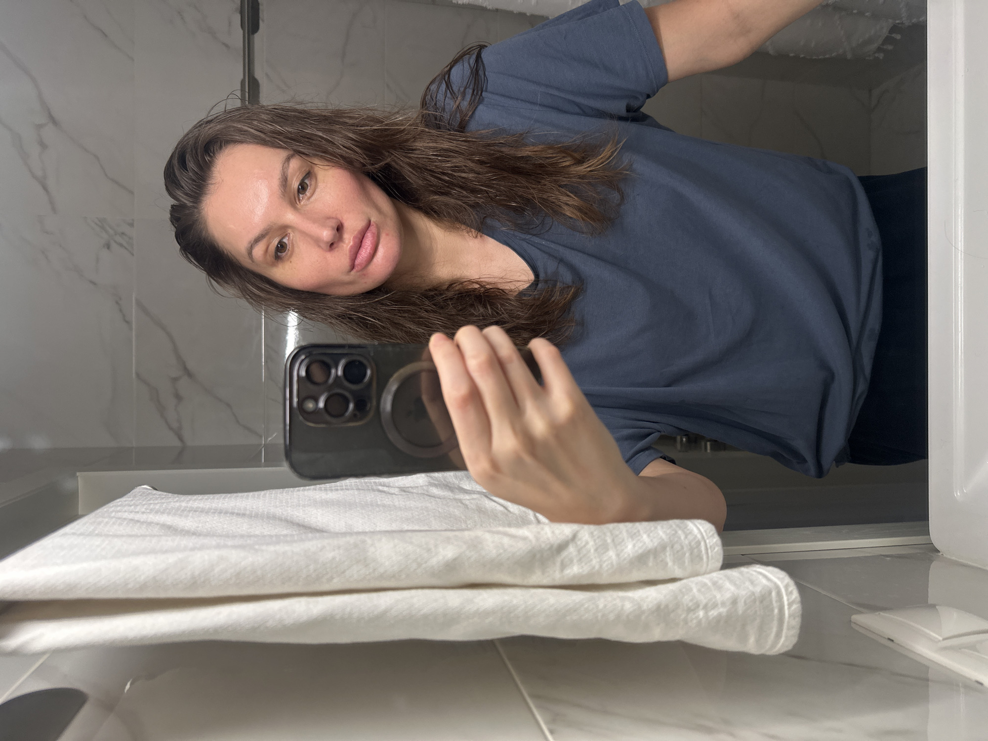 A person is taking a selfie in a bathroom with a marble-tiled wall, a white sink, and a stack of white towels. The person is holding a smartphone. The person is wearing a blue t-shirt. The location is not Moscow, Russia.