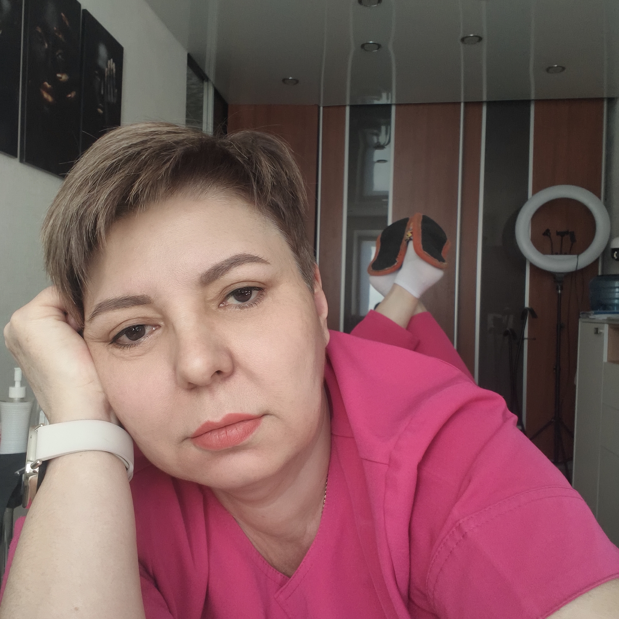 A person with short, light brown hair, wearing a bright pink top and a white smartwatch, is depicted lying prone with her head propped by her left hand. Her gaze is directed at the camera. Her feet, dressed in white socks and dark slippers, are visible bent upwards in the upper right background. The scene is an indoor room in the city of Unknown, Unknown. The background features a light grey wall on the left with two vertical dark-framed art pieces exhibiting abstract yellow-orange forms. A white pump bottle rests on a surface in the lower left. The right background displays a multi-paneled wall or closet system, a white ring light on a black tripod, and a blue water cooler bottle positioned on a white-topped cabinet. The ceiling is light-colored with multiple recessed circular lights.